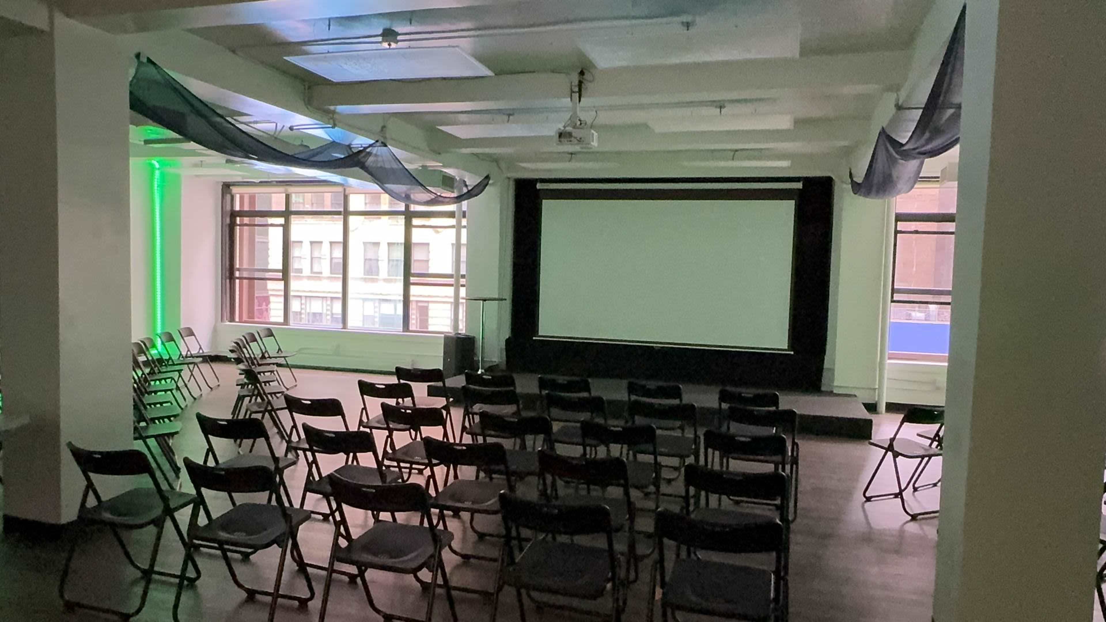 The image shows a spacious indoor room set up for a presentation, featuring rows of black chairs facing a large projection screen.