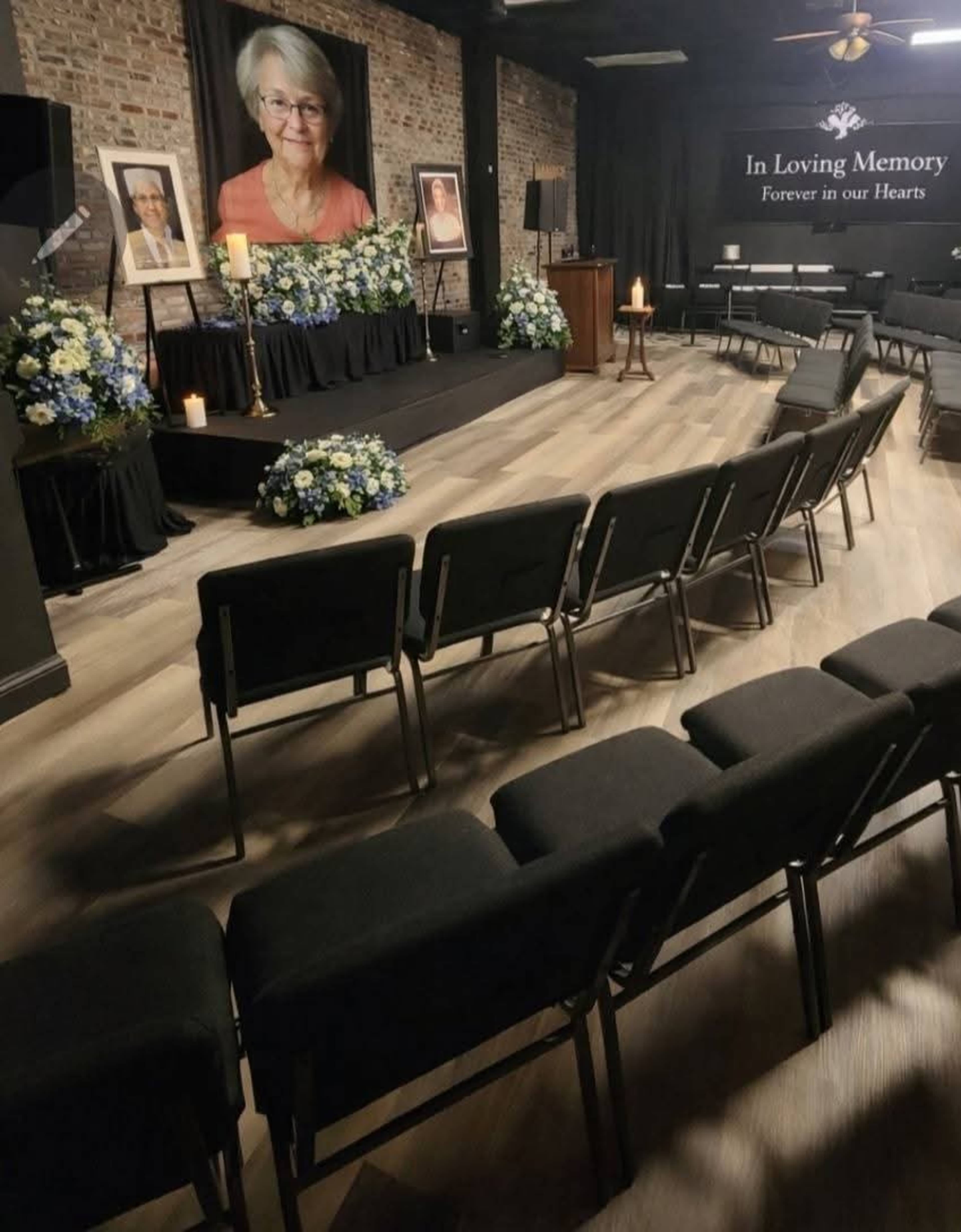 The image shows a memorial service setup with chairs arranged in rows facing a stage adorned with floral arrangements, candles, and framed photographs of a person, against a brick wall backdrop.