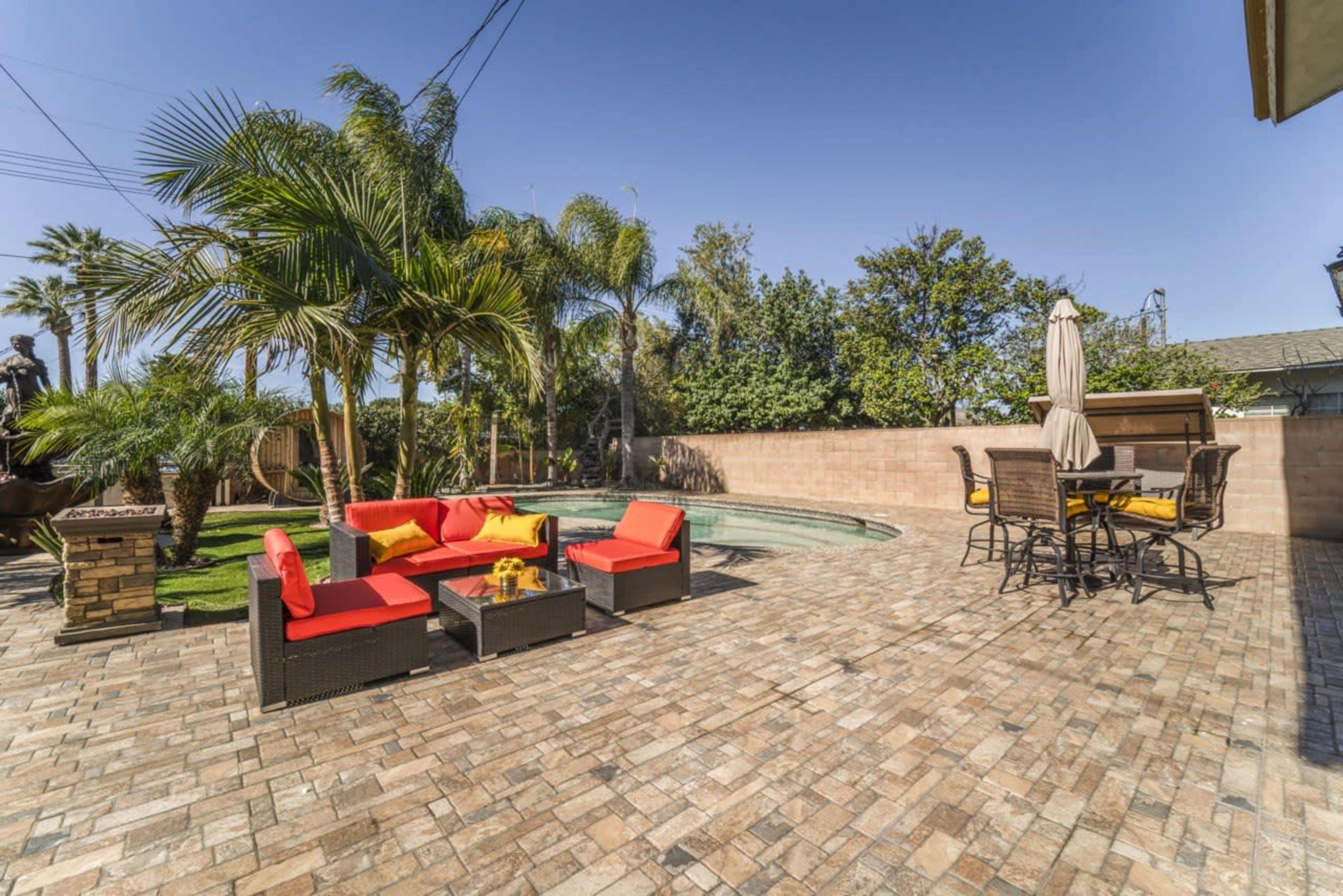 A patio area with red outdoor seating, a table with chairs, a pool, and palm trees in a sunny residential backyard.