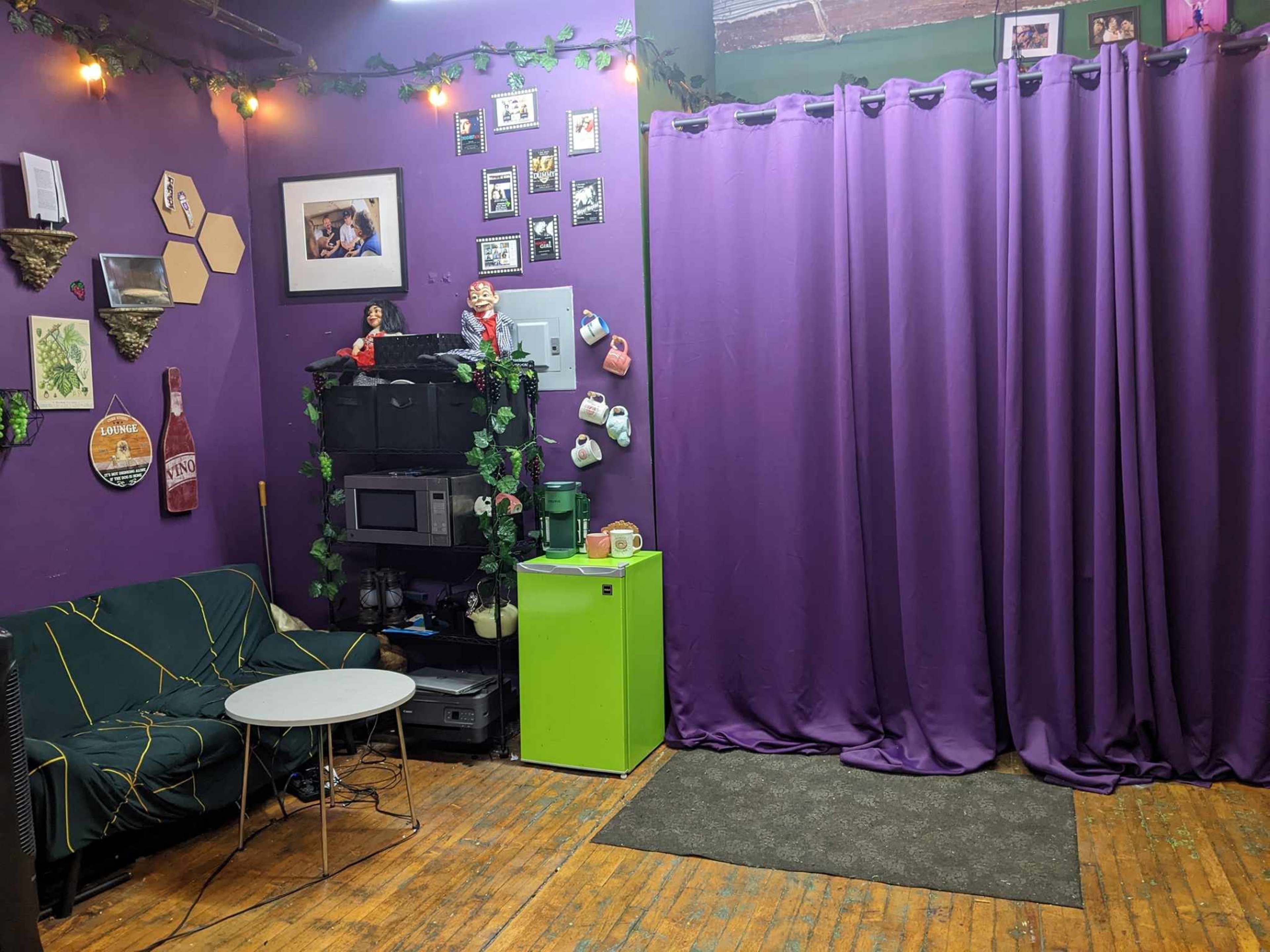 The room features a green refrigerator, a small table, a dark couch, and purple curtains, with photos and decorations on the walls.