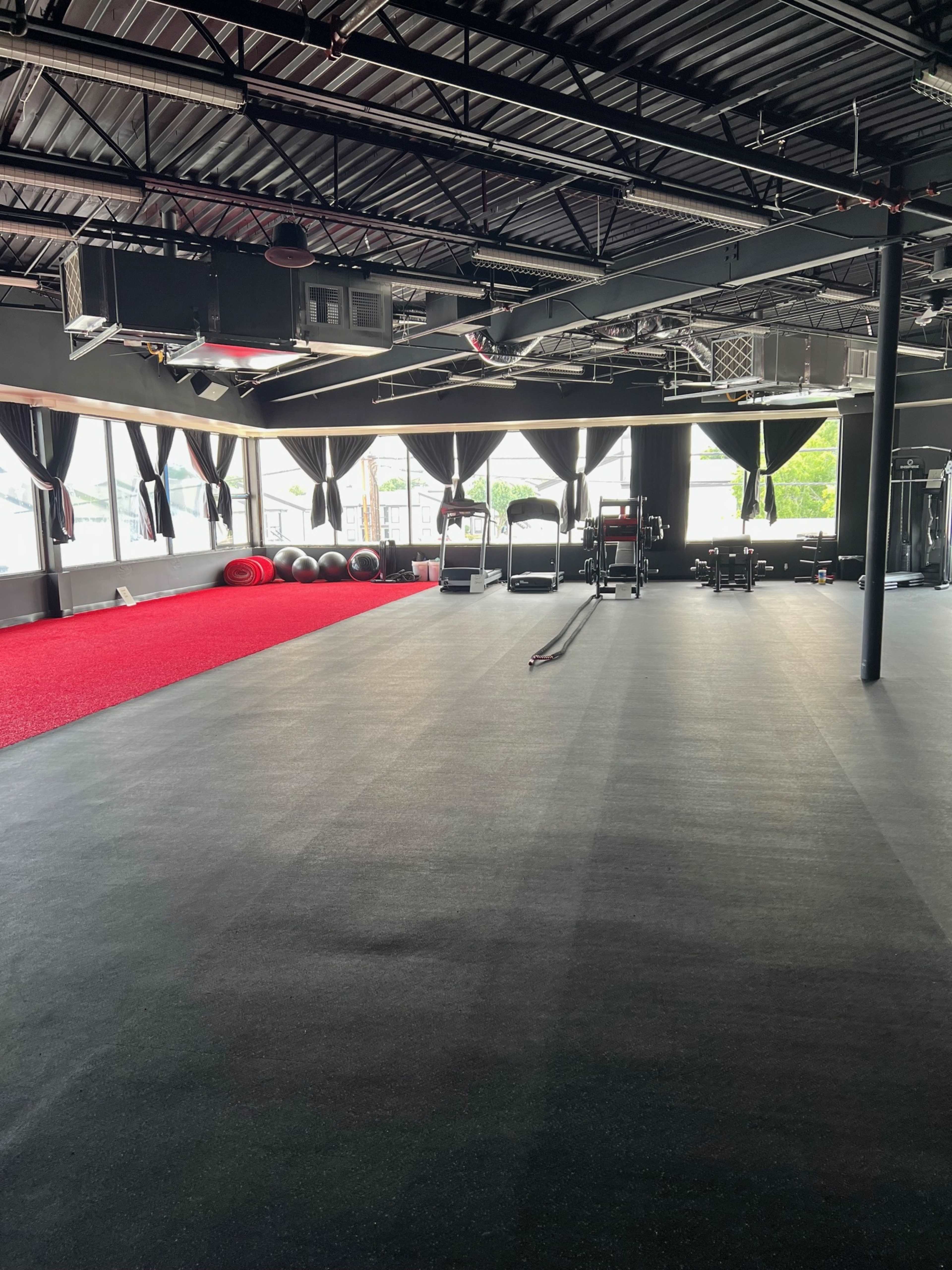 A spacious gym interior with black flooring, exercise equipment, and large windows dressed with dark curtains.