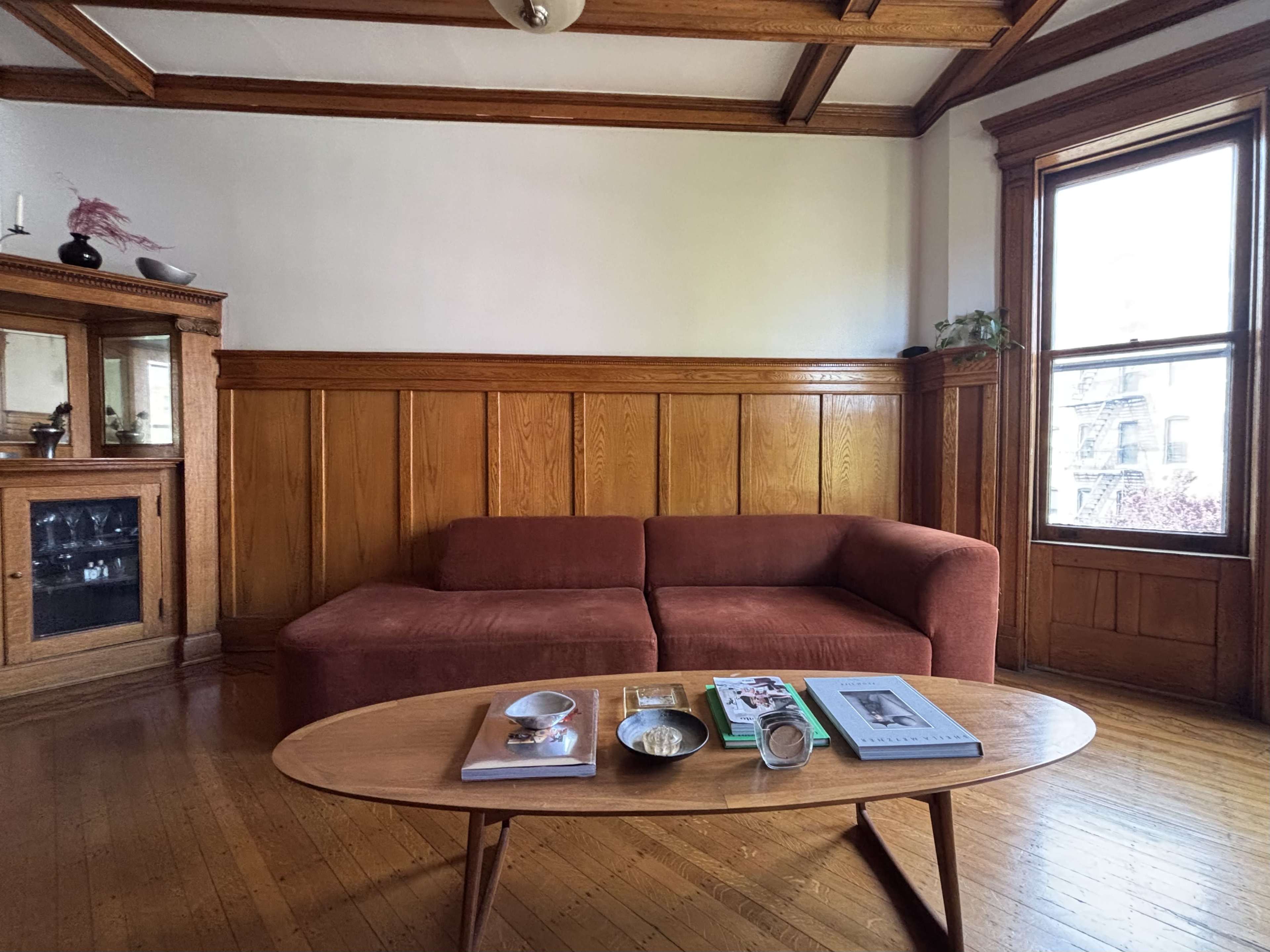 The image shows a living room with a brown sectional sofa, a wooden coffee table with books and decorative items, and wood-paneled walls with a window letting in natural light.
