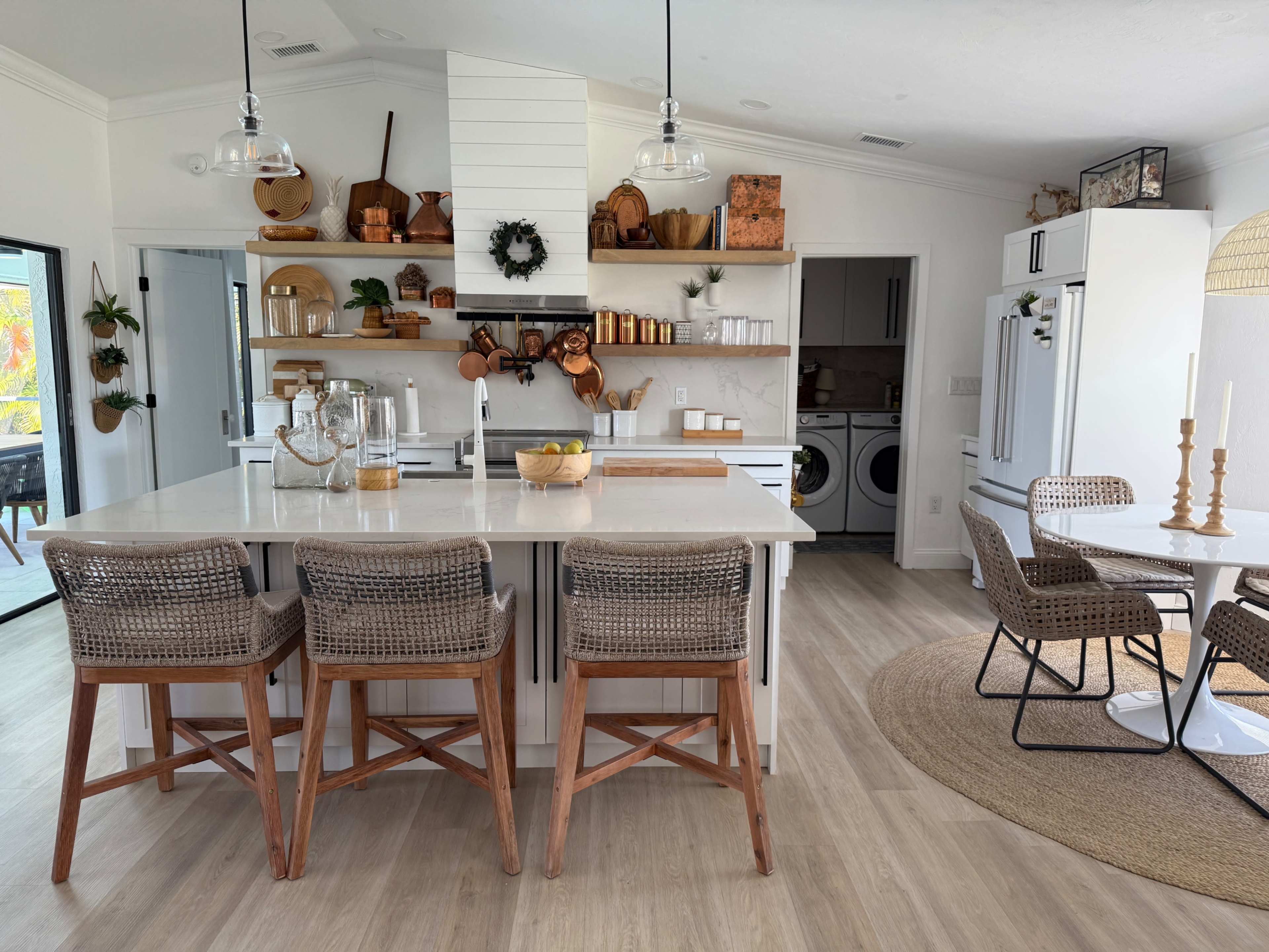 A modern kitchen features a central island with bar stools, open shelving displaying decorative items and kitchenware, and a dining area with a round table and woven chairs.