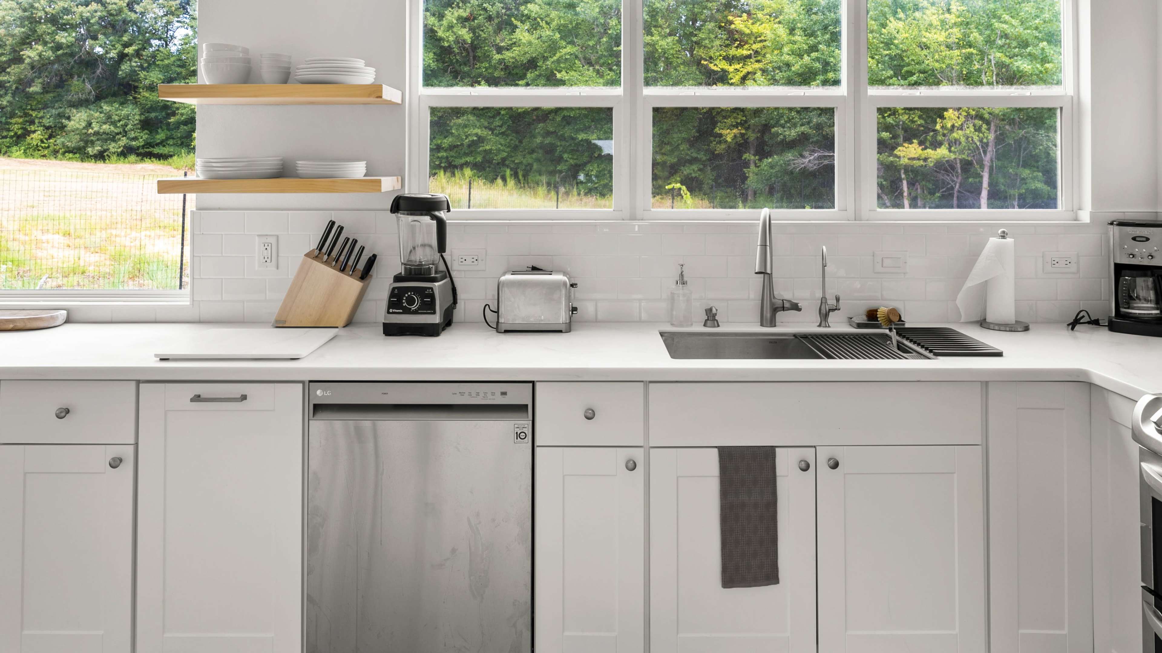 A modern kitchen features white cabinetry, a stainless steel dishwasher, and a counter with a blender and toaster beside large windows overlooking greenery.