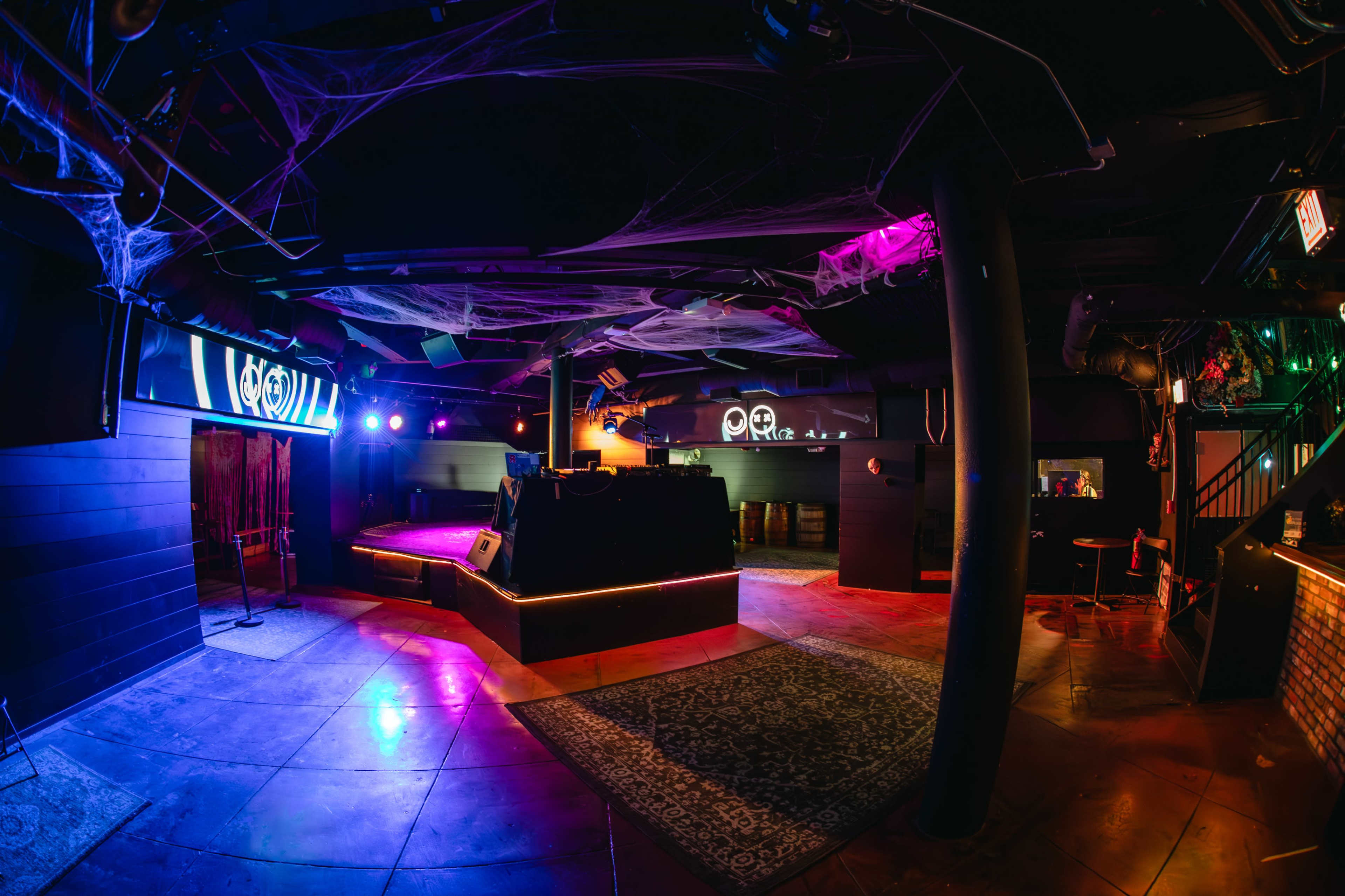 The image shows a dimly lit nightclub interior with colorful lighting, a DJ booth, and decor that includes spiderwebs.