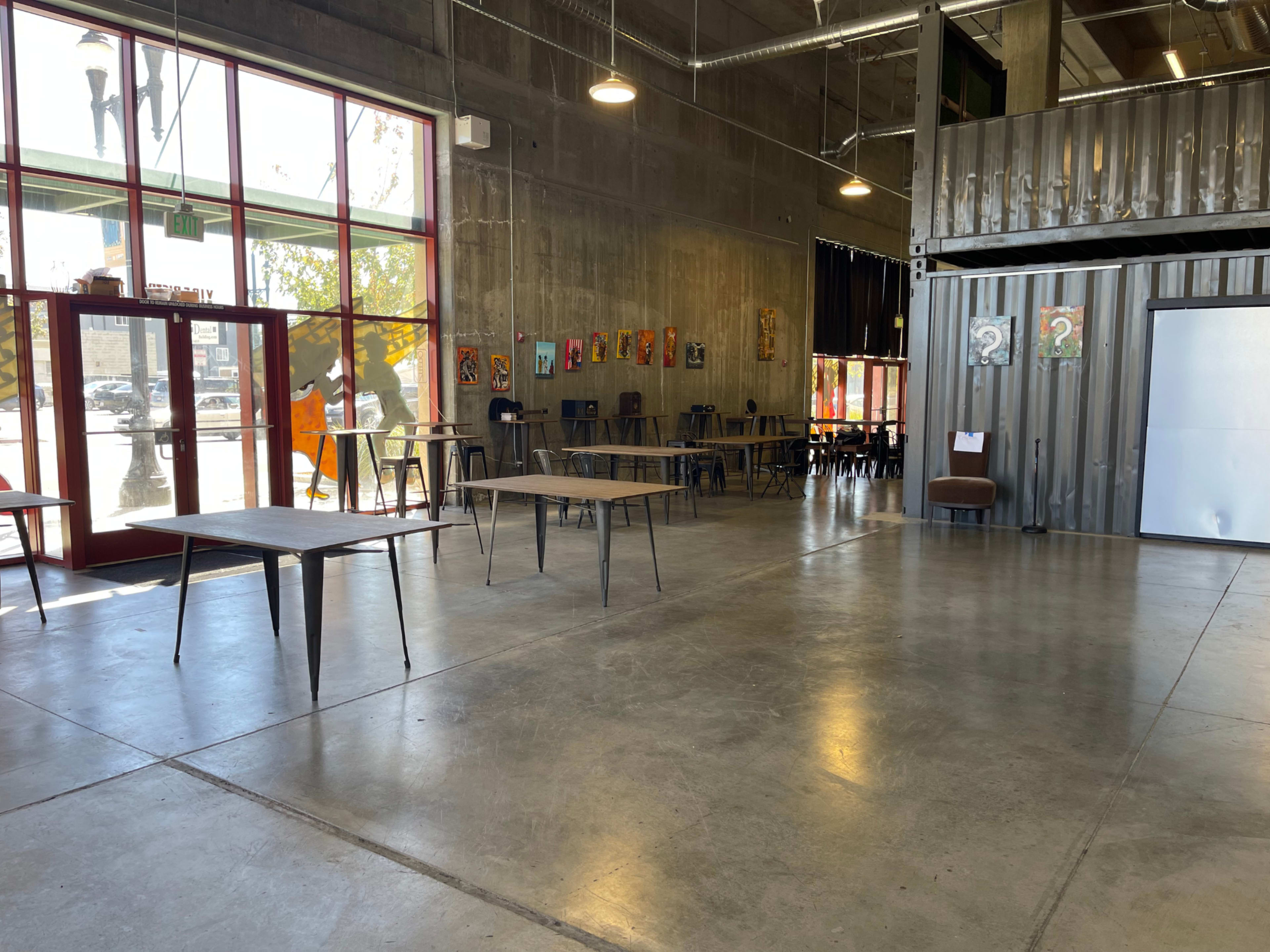 The image shows an industrial-style indoor space with concrete floors, large windows, empty tables, and colorful artwork on the walls.