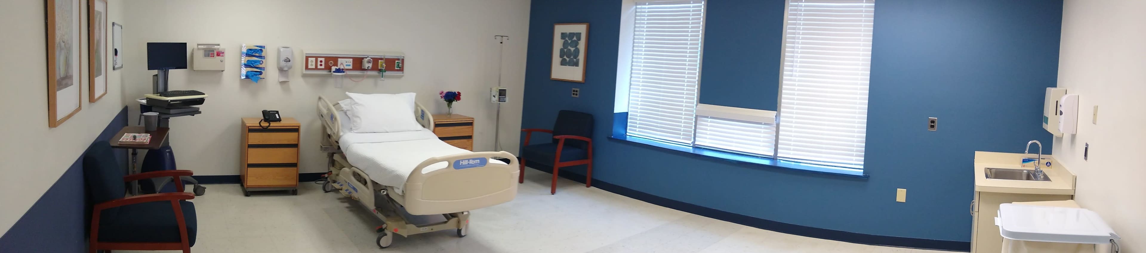 A hospital room with a bed, a wooden bedside table, a chair, and a small sink in a well-lit setting.