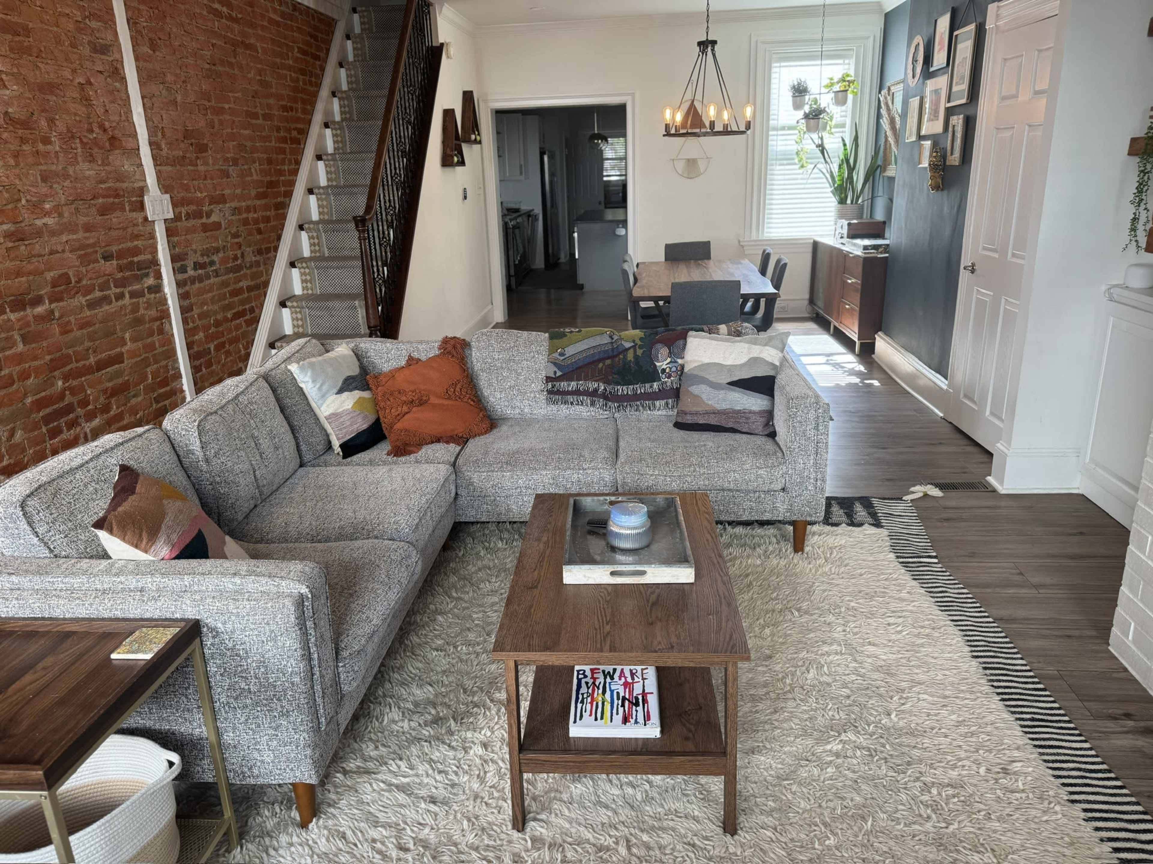The image shows a cozy living room with a grey sectional sofa, a wooden coffee table, and an area rug, featuring exposed brick walls and access to a dining area in the background.