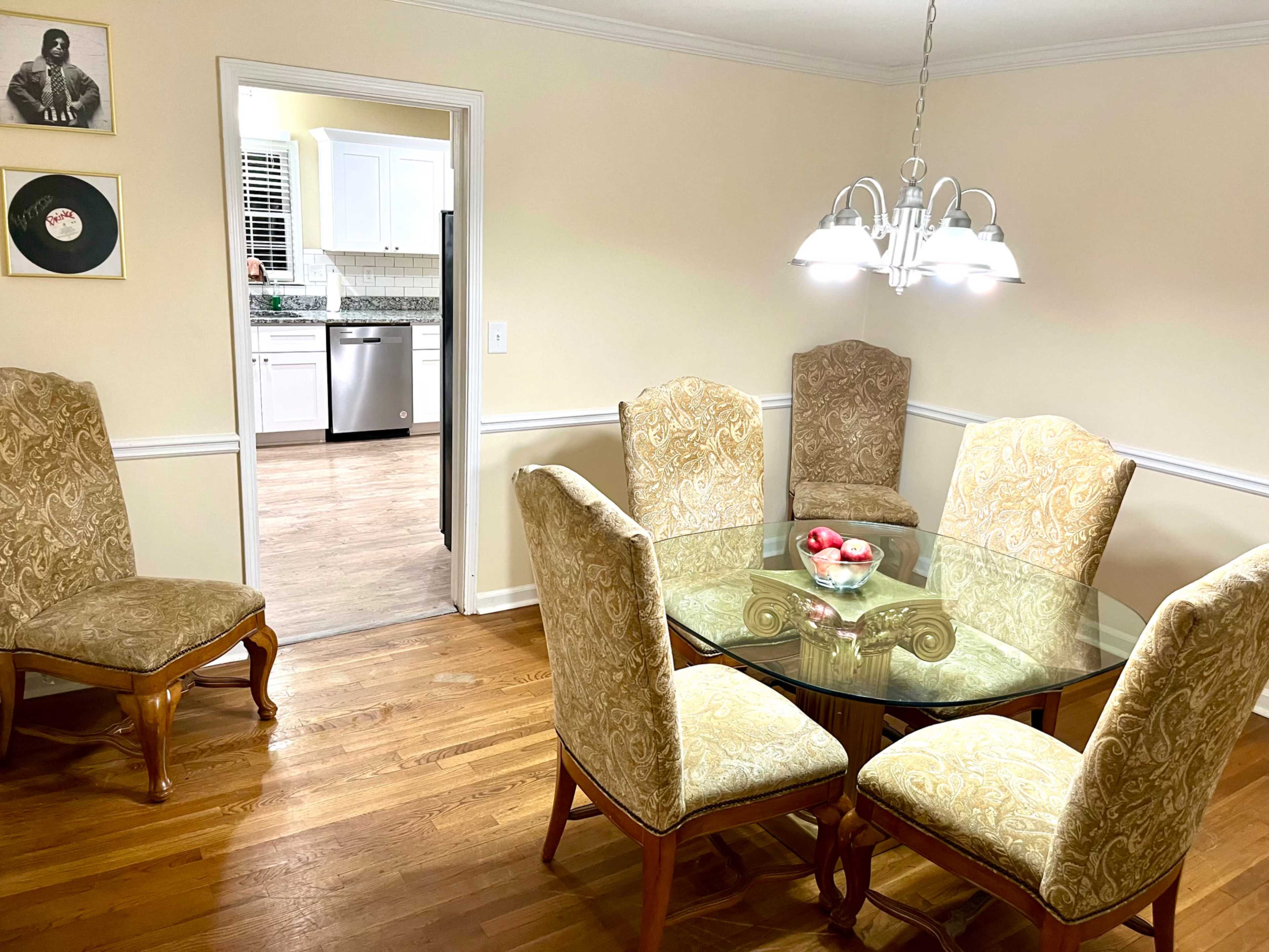 A round glass dining table with six upholstered chairs is set in a brightly lit dining area adjacent to a kitchen.