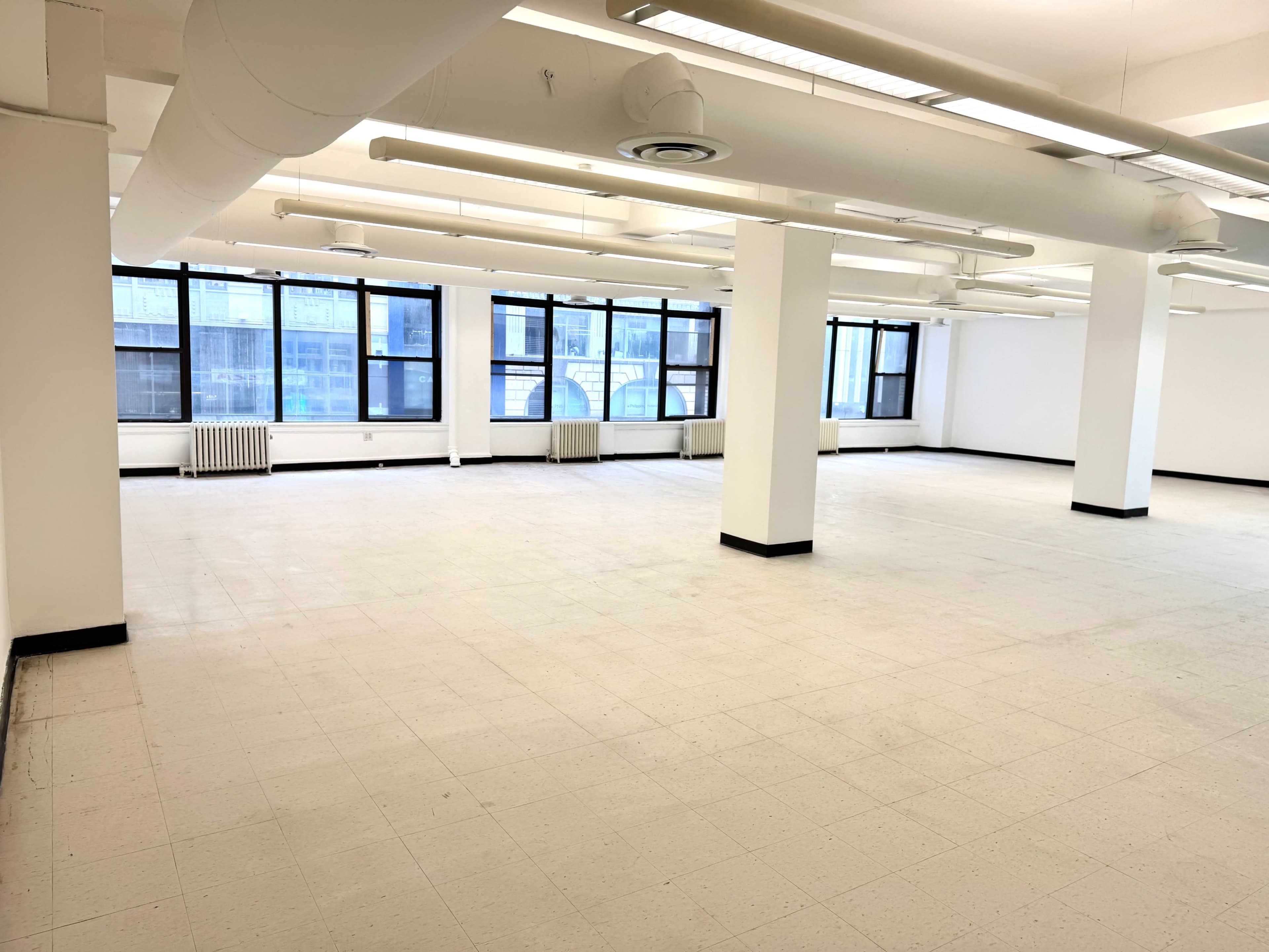 The image shows a bright, empty room with white walls, large windows, and a tile floor.