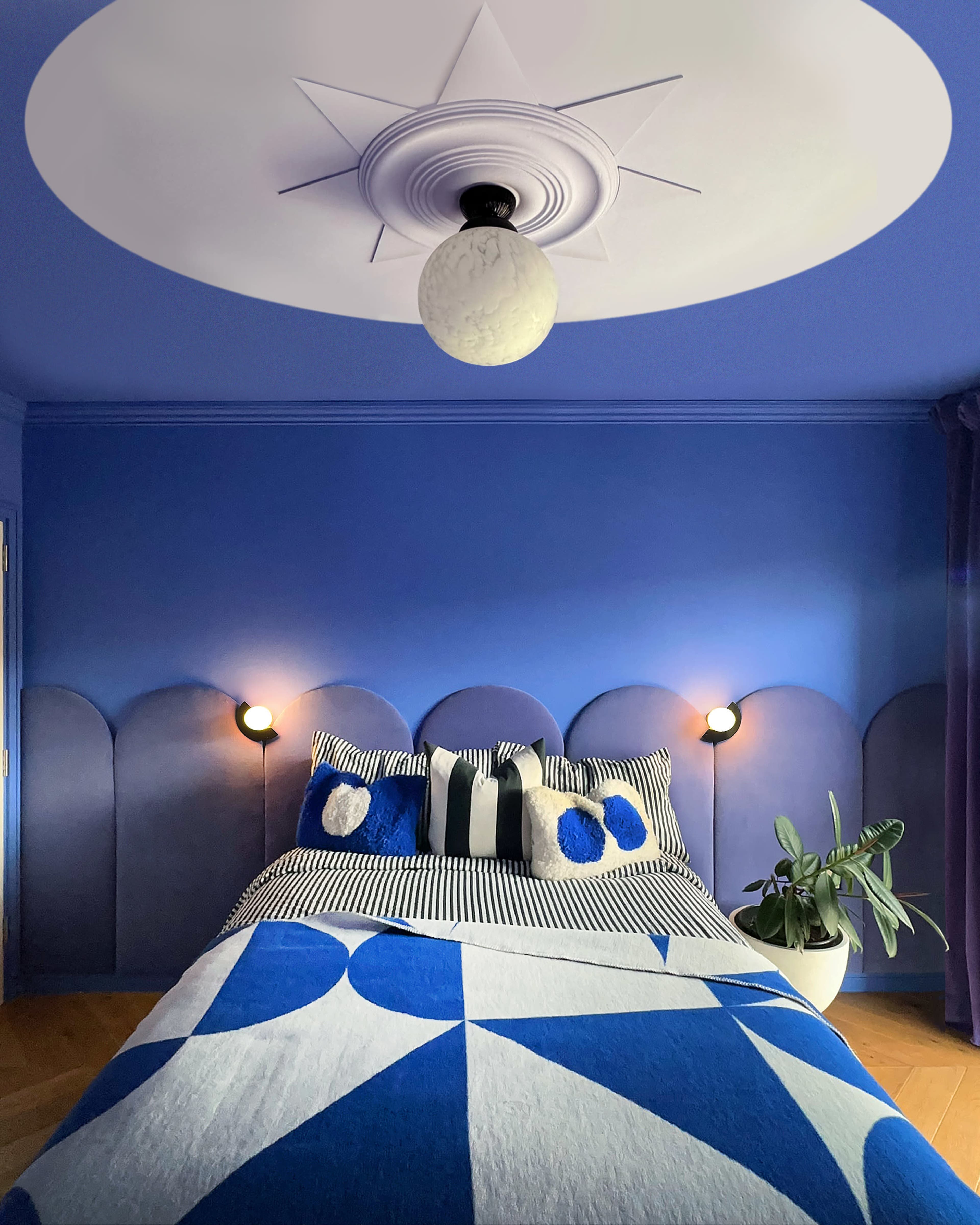 The bedroom features a blue color scheme with a patterned bedspread, a round ceiling light, and wall-mounted lamps beside the bed.