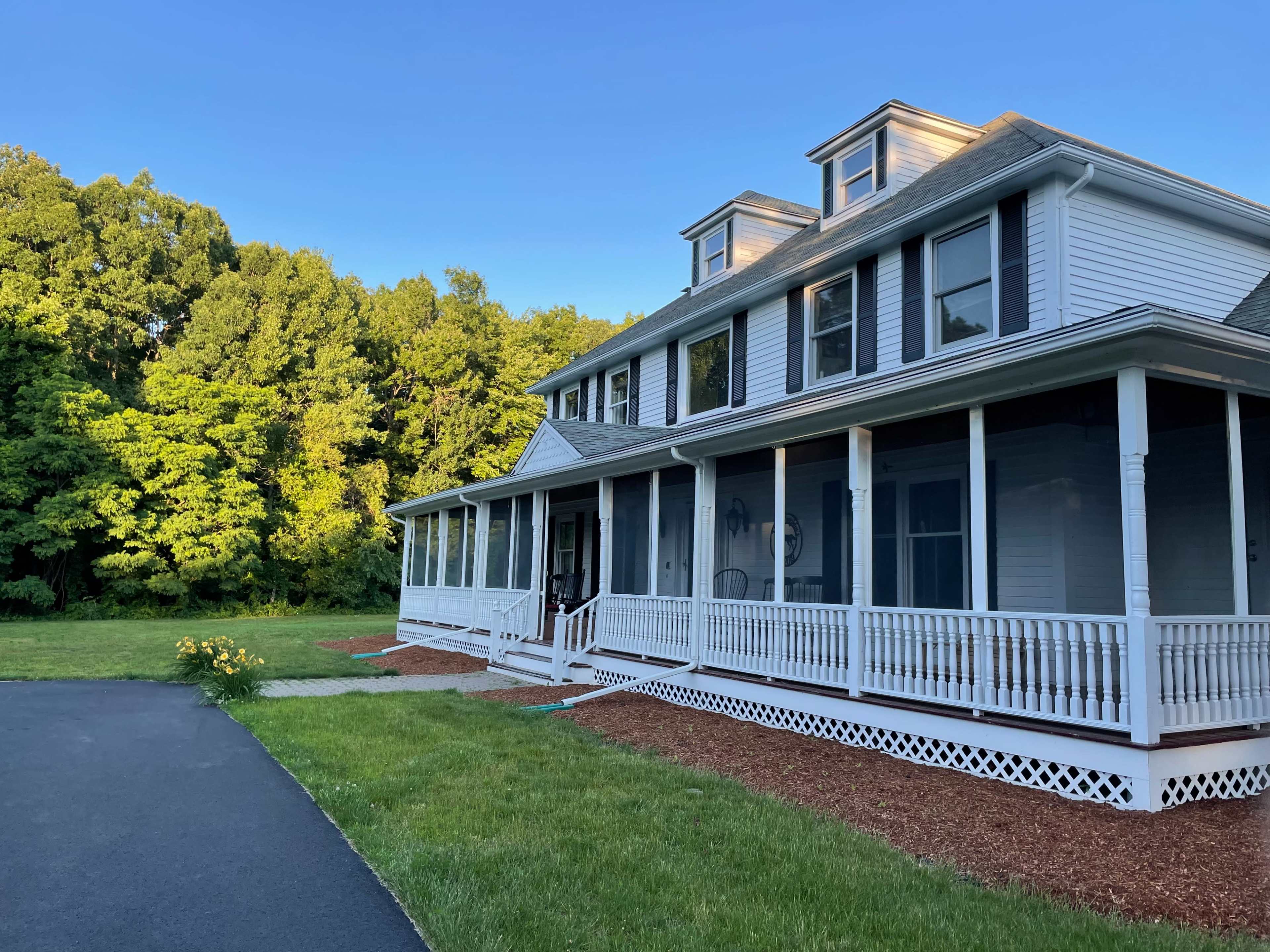 A large, two-story house with a porch and a gravel path sits next to a well-maintained lawn and wooded area.