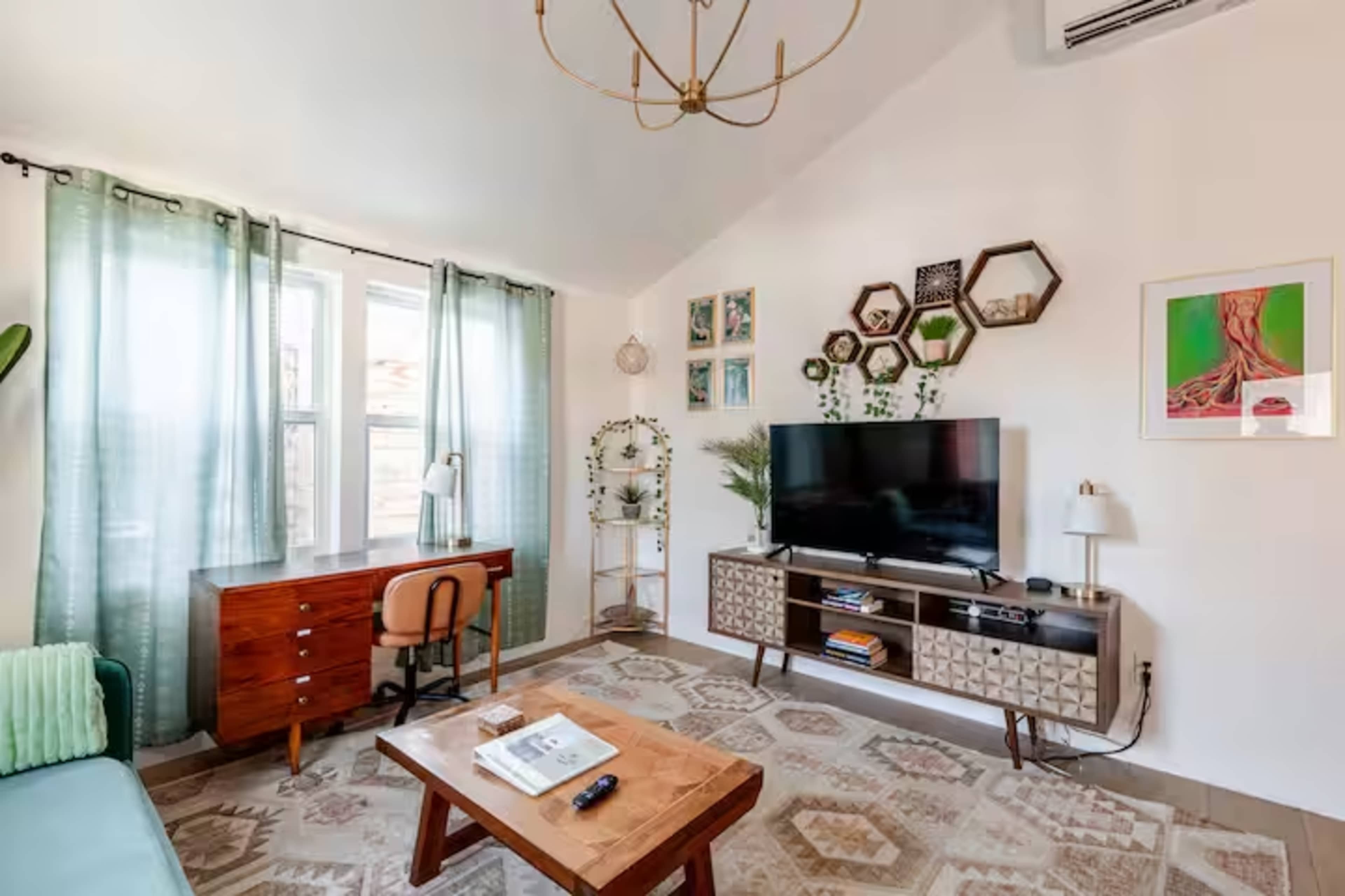 The living room features a television mounted on a cabinet, a wooden desk with a chair, large windows dressed with sheer curtains, and various decorative elements on the walls.