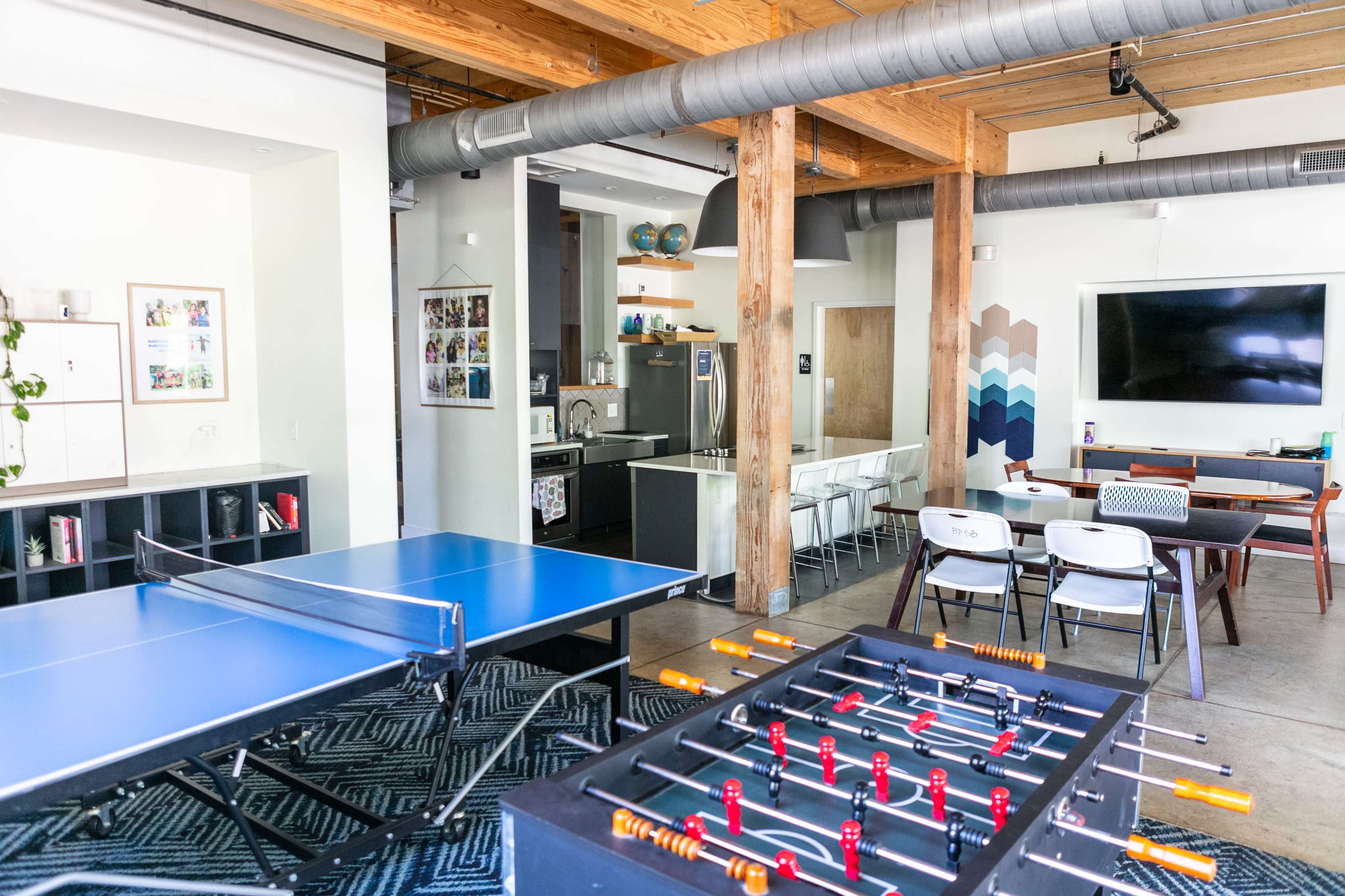 The image shows a spacious room featuring a ping pong table, a foosball table, a dining area with white chairs, and a large television mounted on the wall.