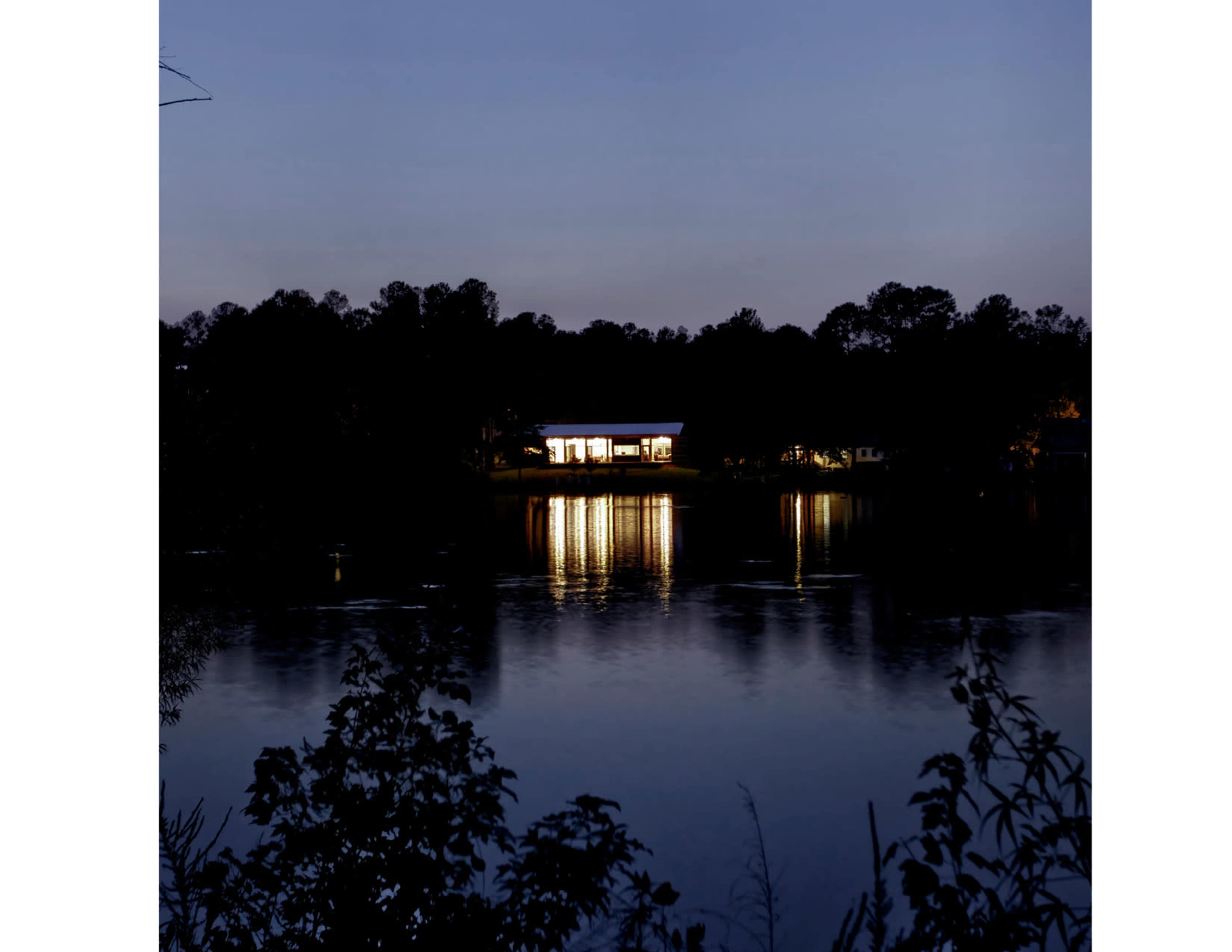 A modern building is illuminated along the shoreline of a calm river at dusk.