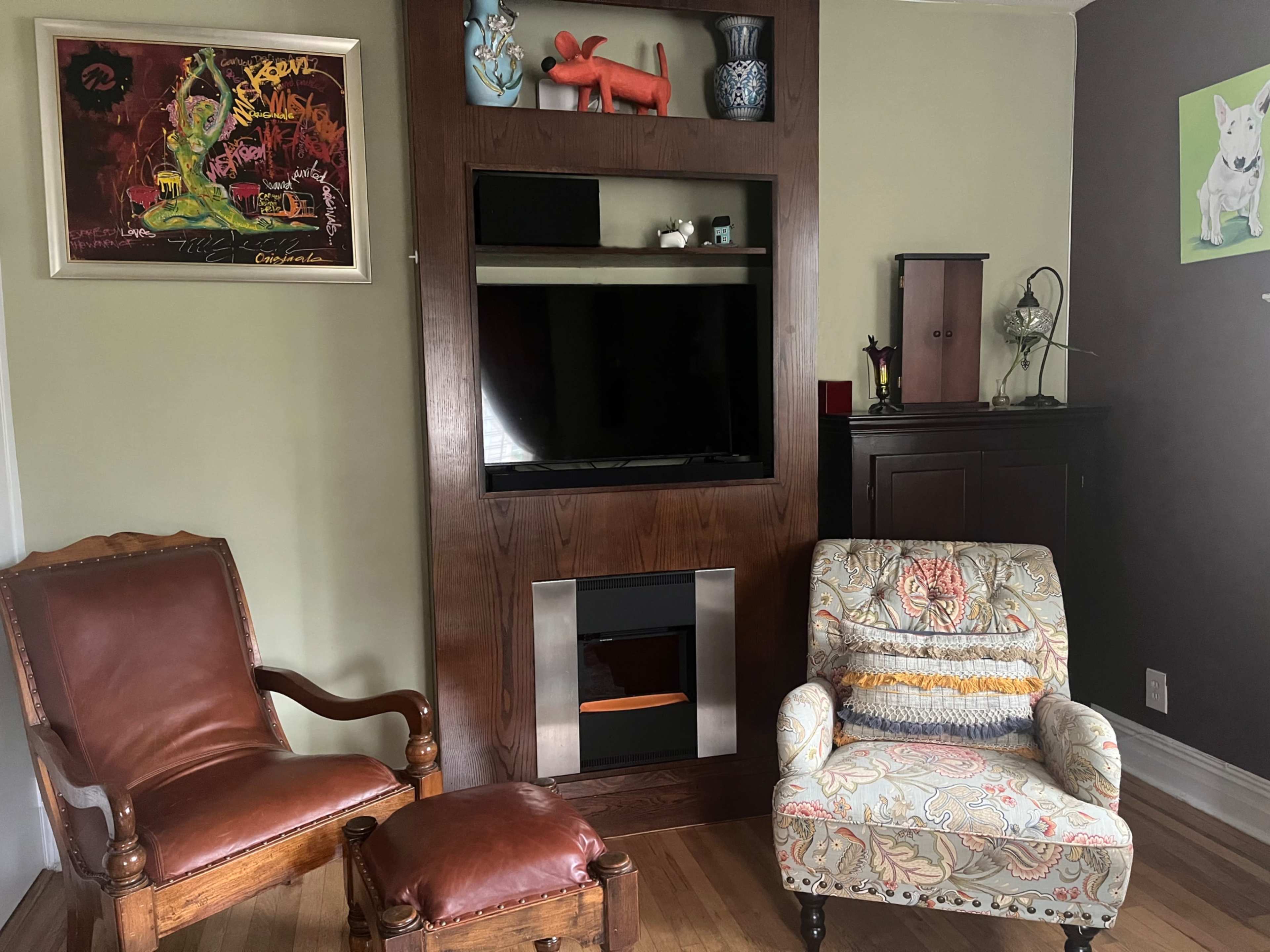 The image shows a living room with two armchairs, a television mounted in a wooden cabinet, and decorative items on the shelves.