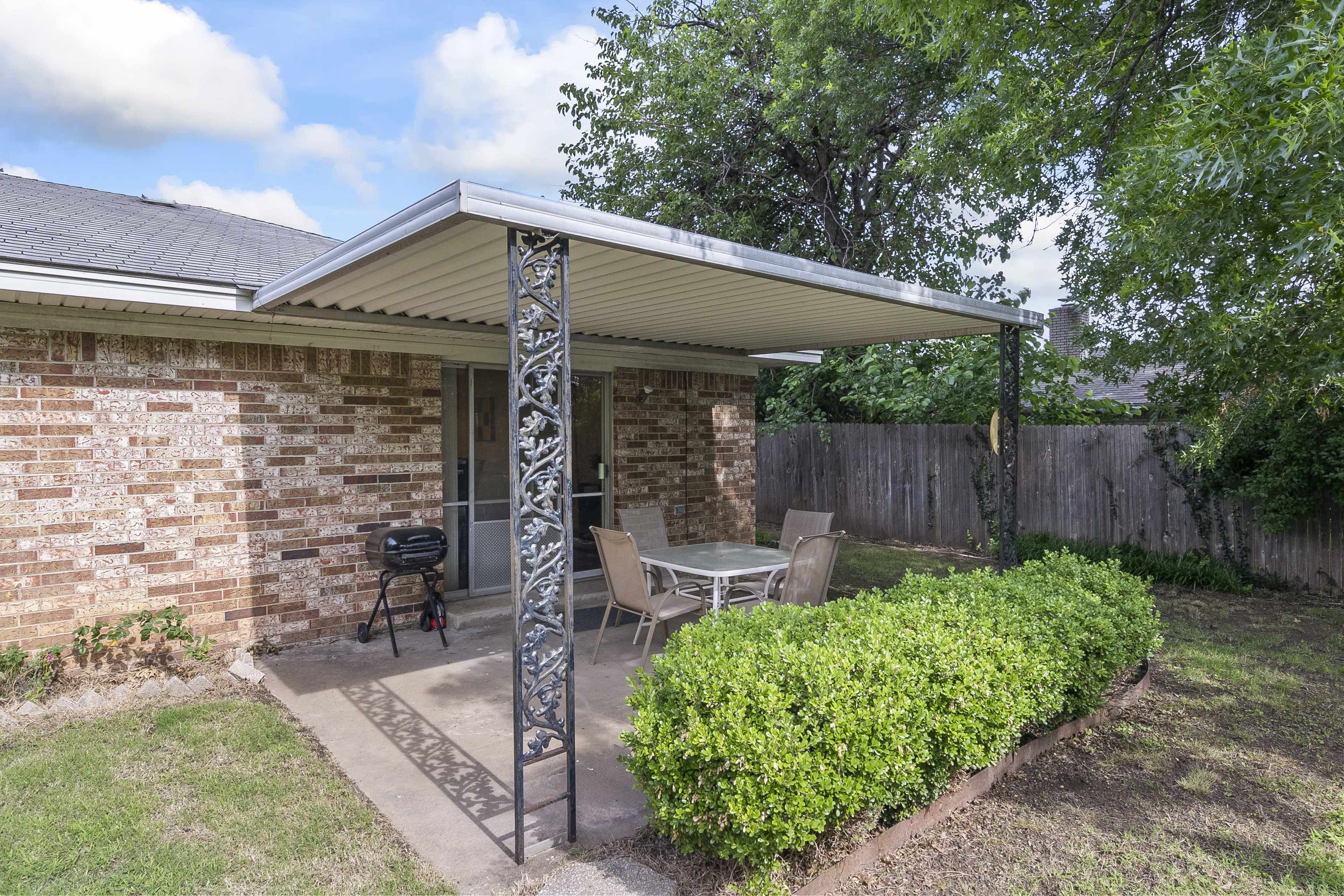 A patio area features a metal canopy with a table and chairs, bordered by a small hedge and a grill nearby, set against a brick wall and landscaped yard.