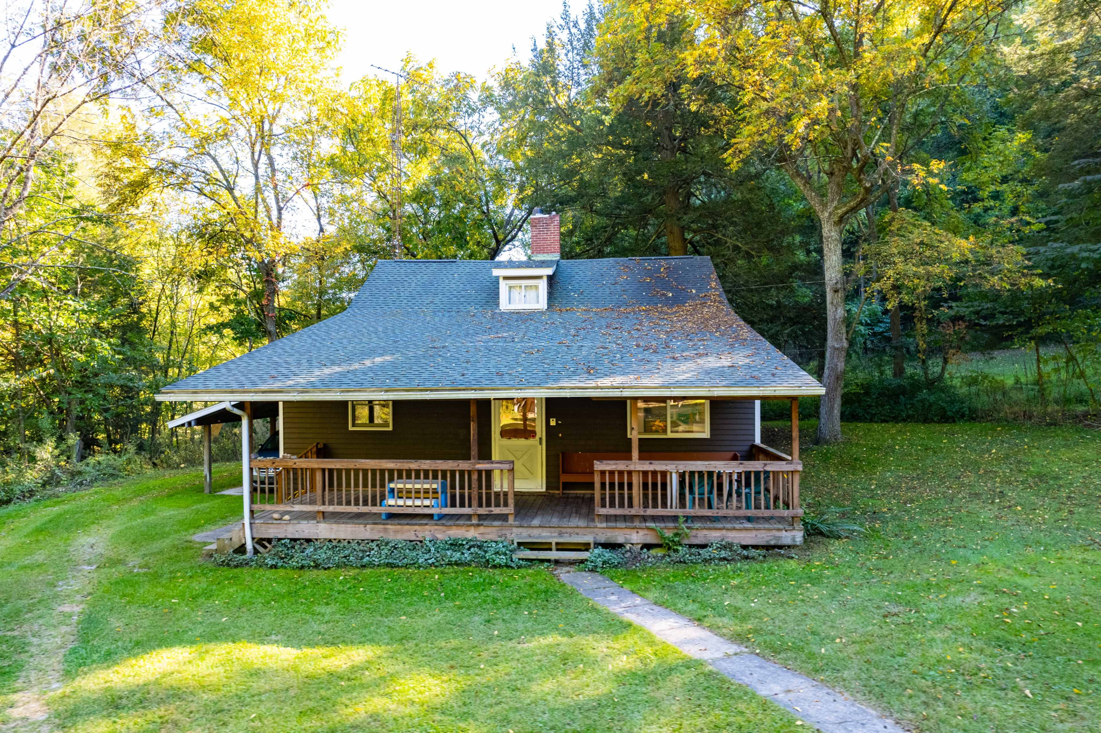 A rustic house with a sloped roof and a front porch is surrounded by trees and a grassy lawn.