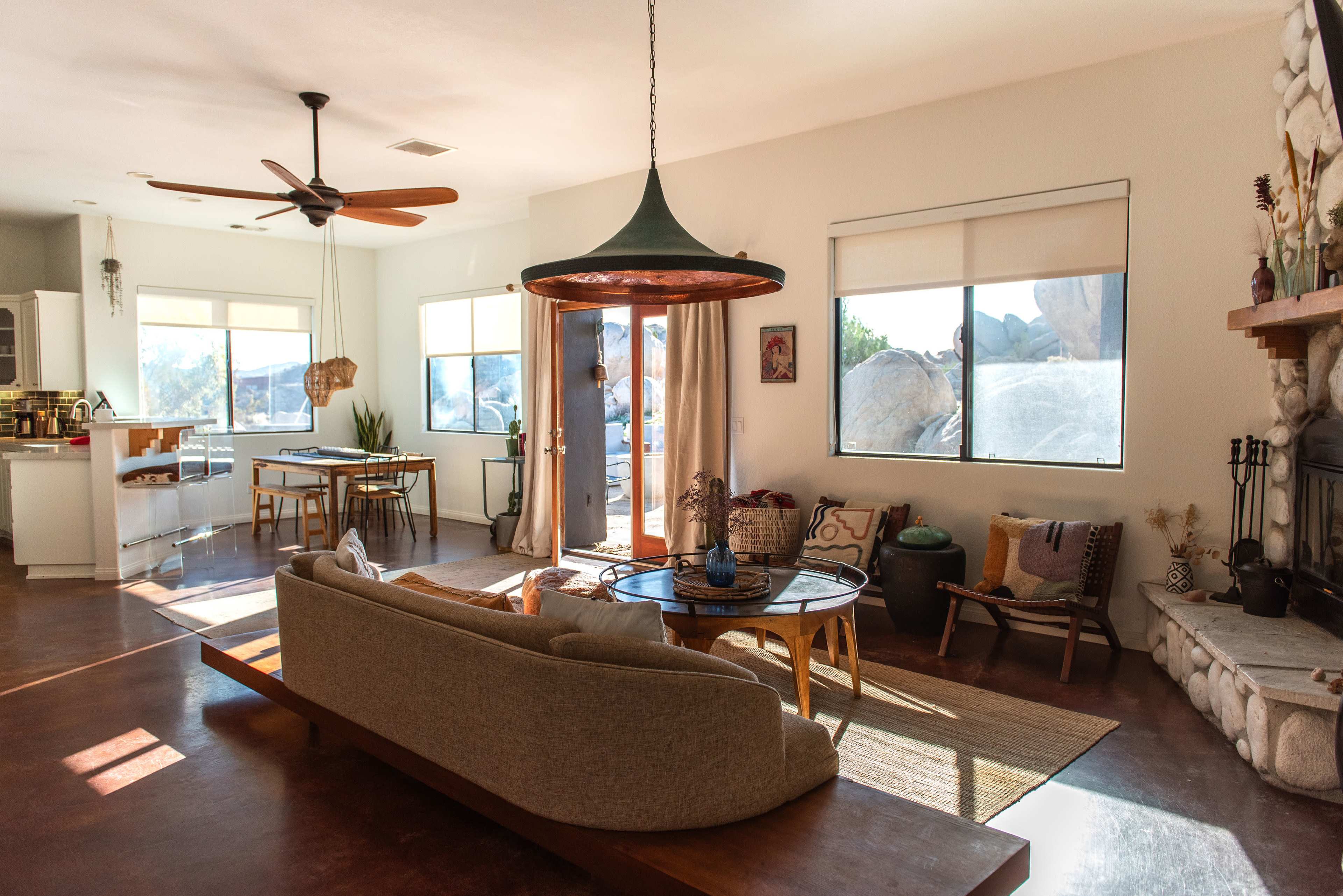 The image shows a spacious living room with a large sofa, a circular coffee table, and large windows letting in natural light, with a modern kitchen visible in the background.