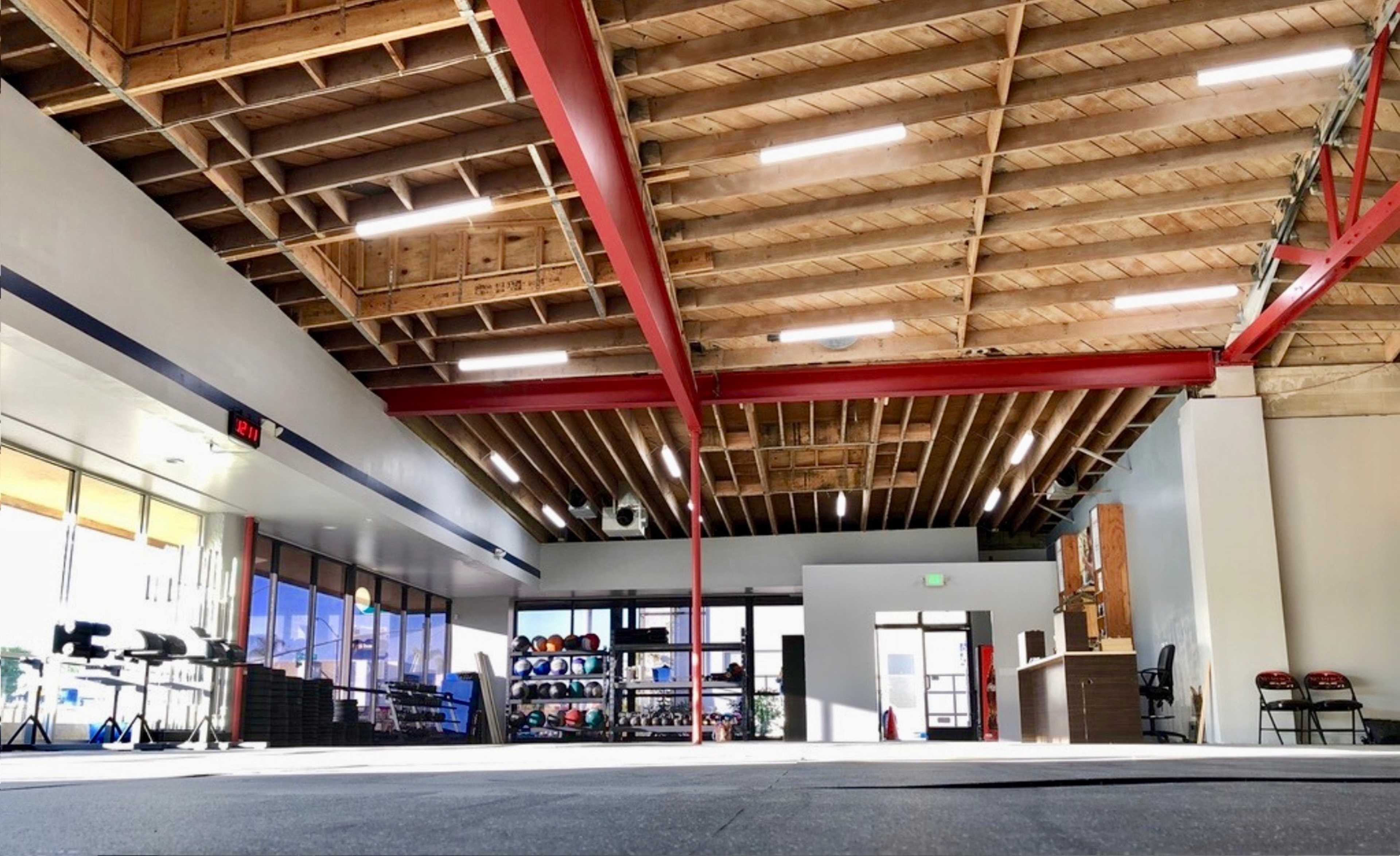 The image shows a spacious gym interior with exposed wooden beams, fitness equipment along the walls, and a large open area with a black rubber floor.