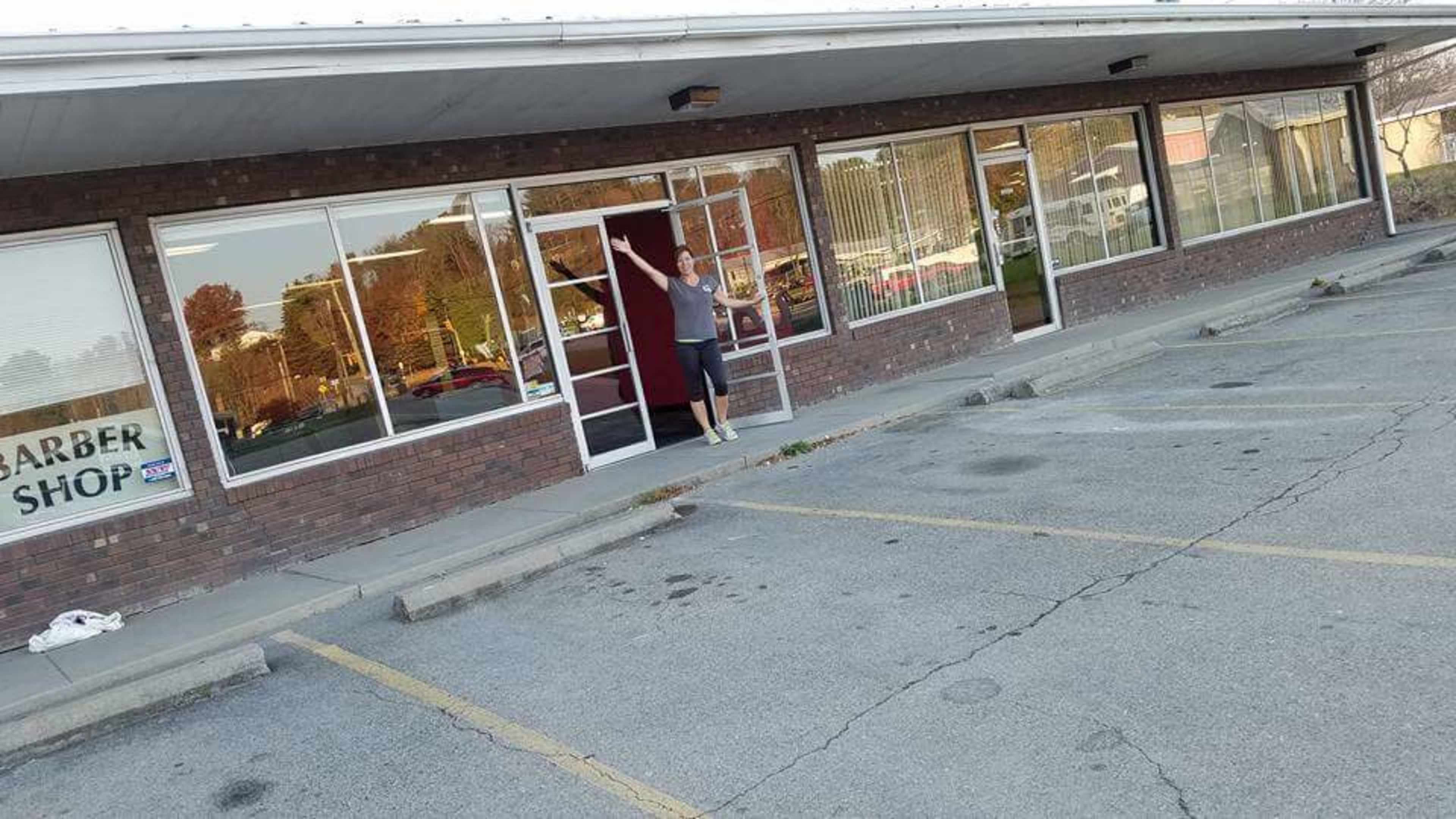 A man stands in front of a barber shop with large glass windows, showcasing a parking lot and surrounding buildings.