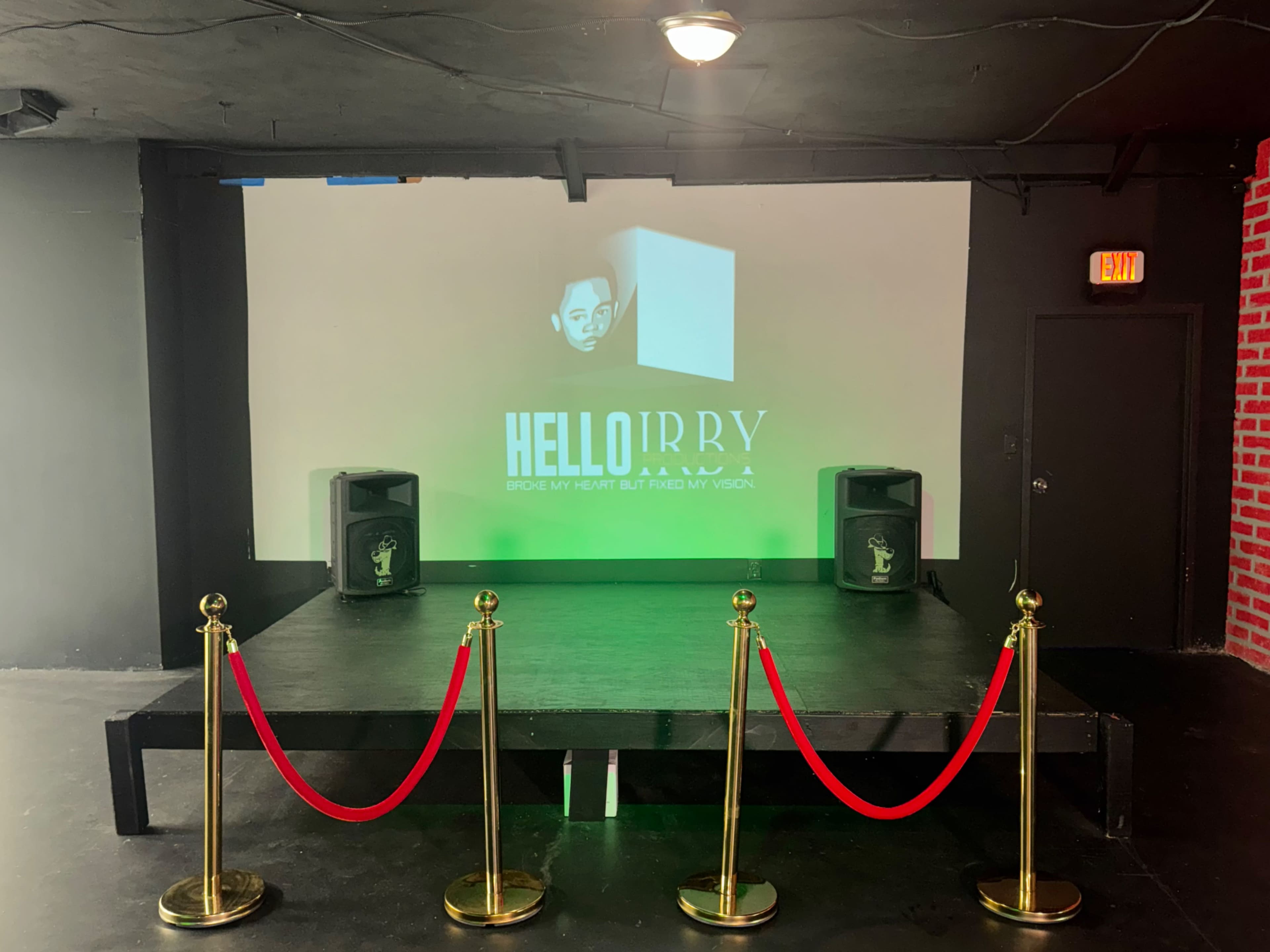 The image shows a stage with a white projection screen displaying a logo, flanked by two speakers and red velvet ropes in front.