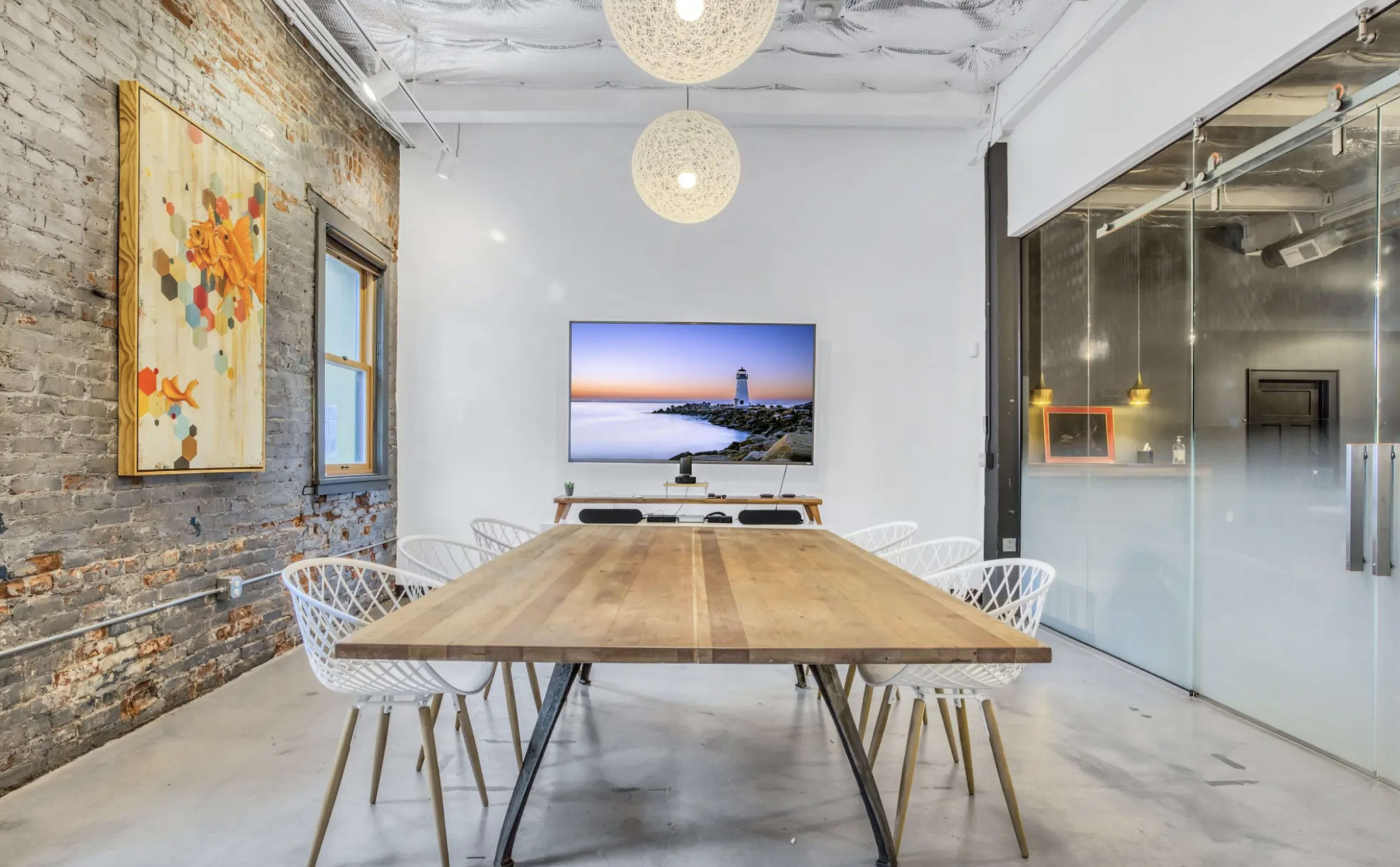 A modern conference room features a large wooden table surrounded by white chairs, with a television mounted on the wall displaying a coastal scene and a glass partition leading to another area.
