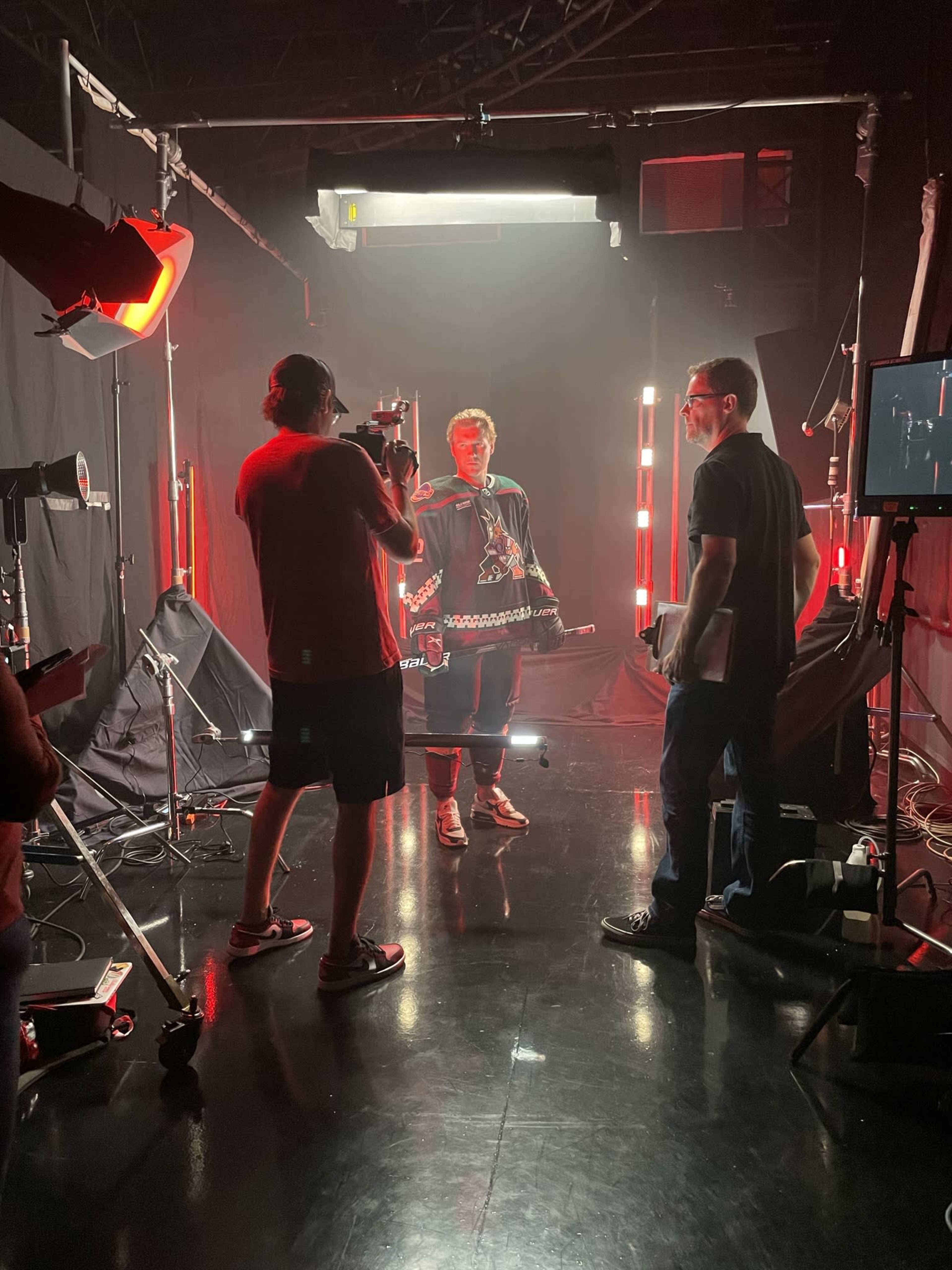 A person in a dark outfit stands in a studio surrounded by bright red lights while crew members operate cameras and equipment.