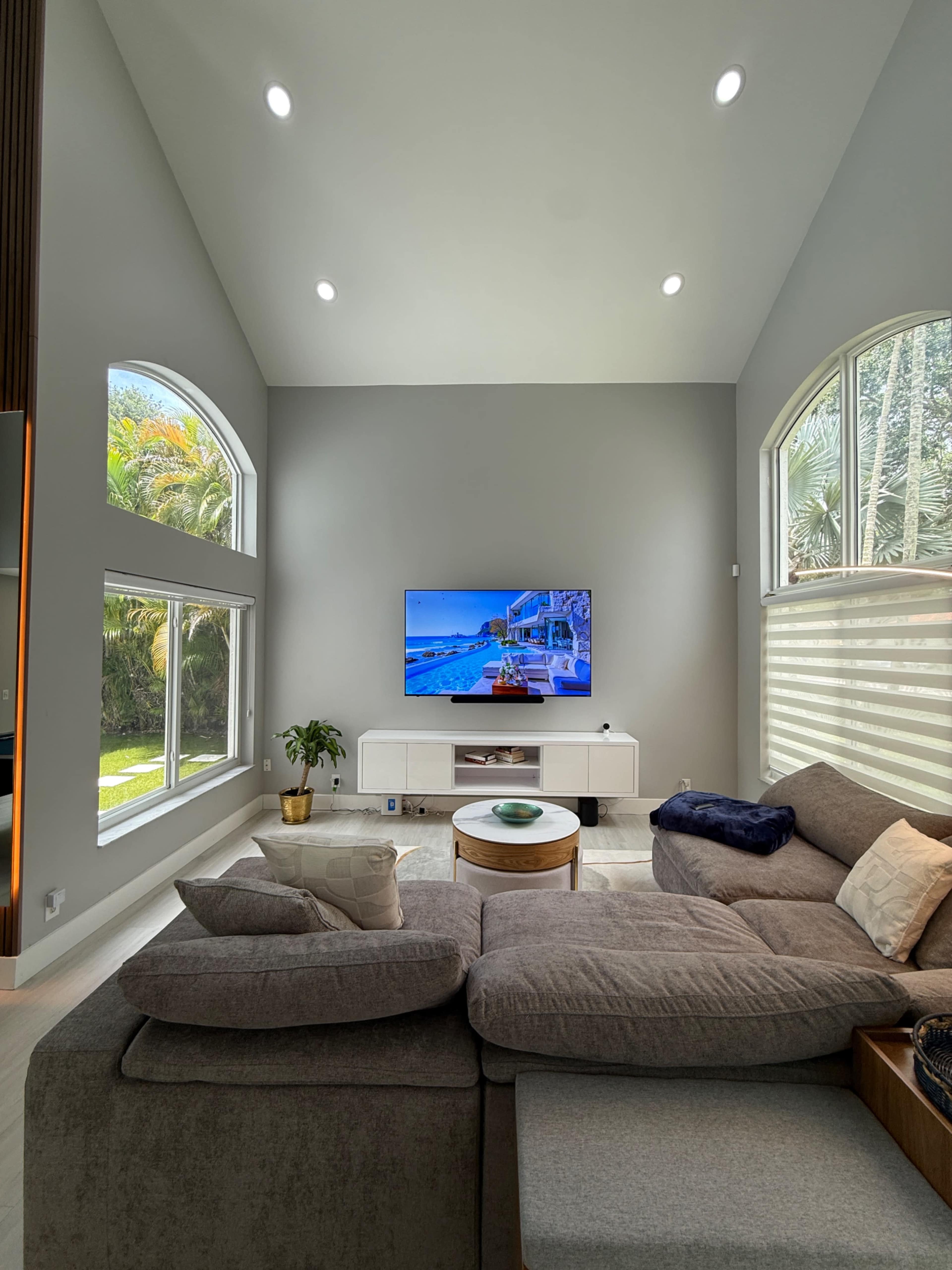 A modern living room features a large sectional sofa facing a wall-mounted television, with windows allowing natural light and greenery visible outside.