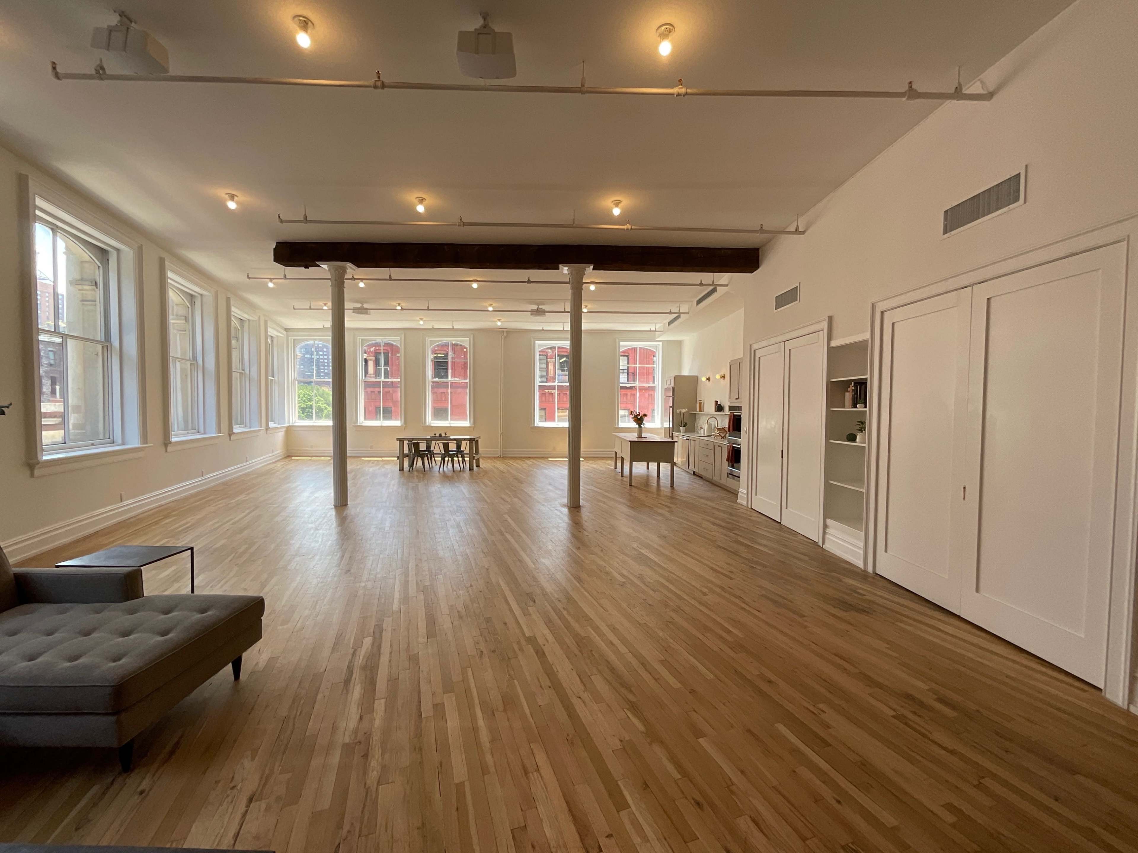 A spacious, well-lit room with wooden floors, large windows, a dining table, and a kitchenette in the corner.