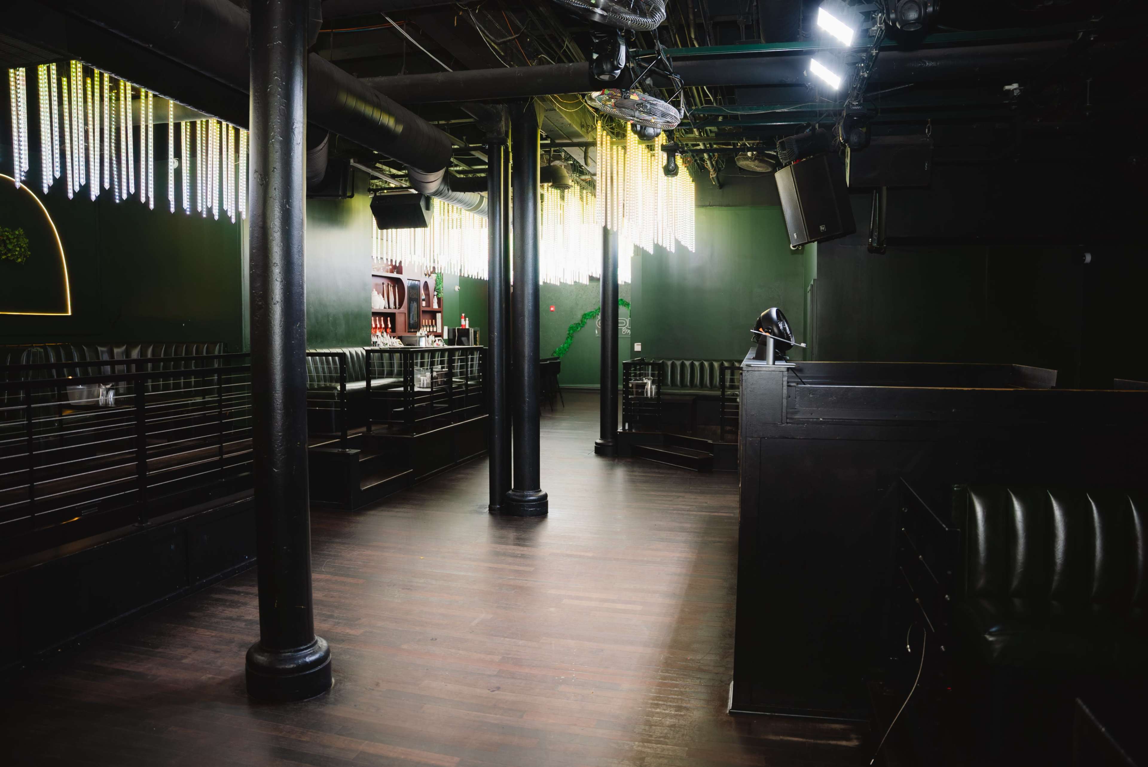 The image shows a dimly lit interior of a club or lounge, featuring dark wooden floors, black furnishings, and a bar area illuminated by hanging light fixtures.