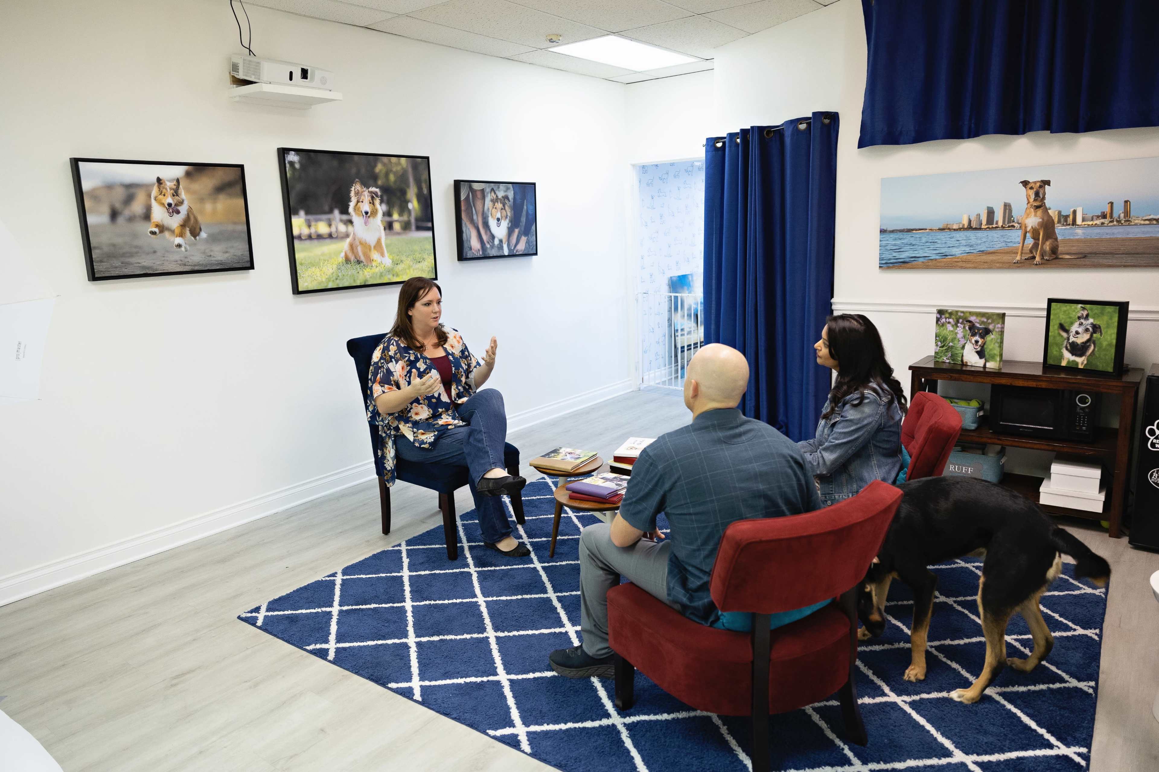 A woman gestures while speaking to a couple seated on red chairs in an office decorated with dog photographs.