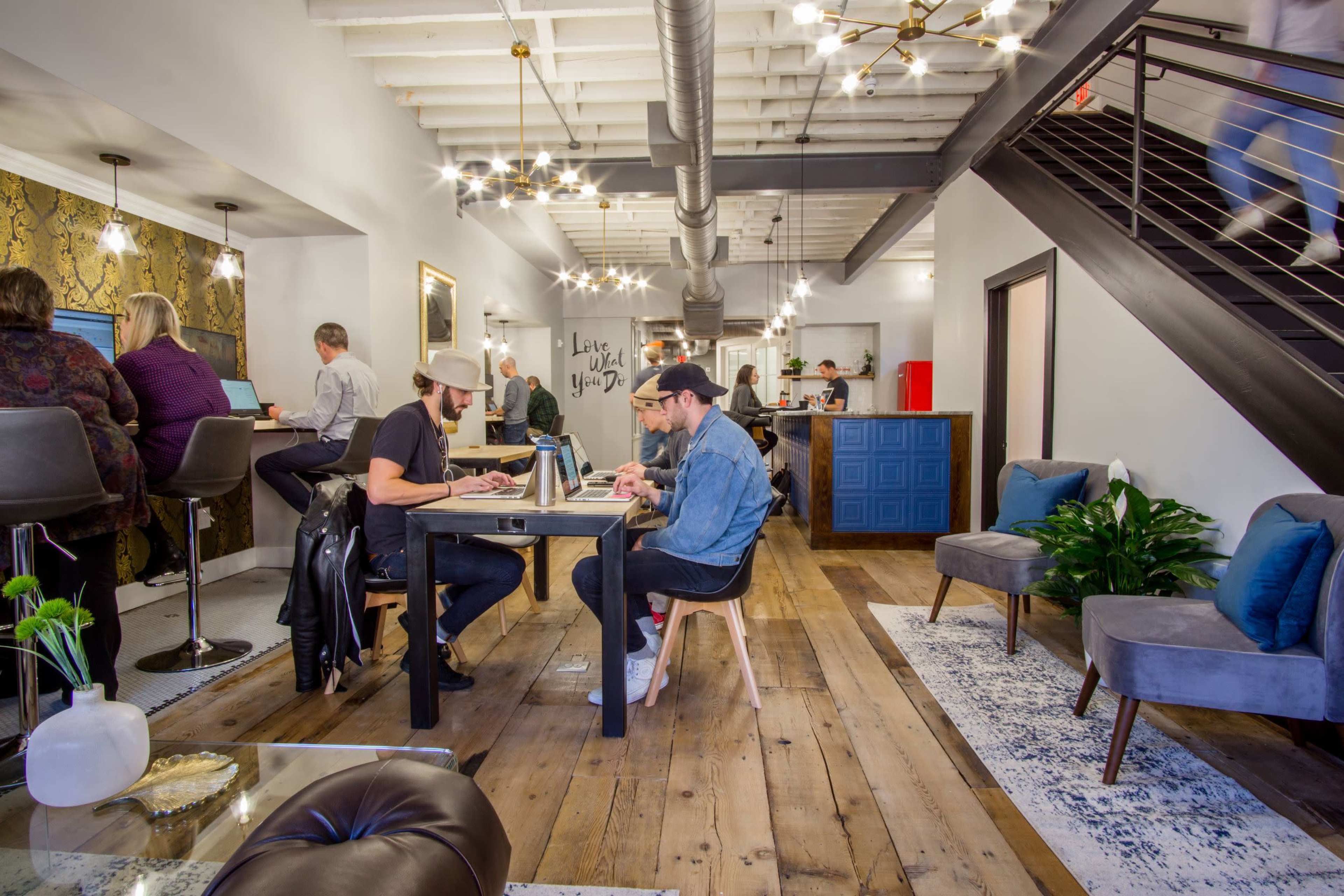 A modern coworking space features several individuals working at tables and a staircase leading to an upper level.