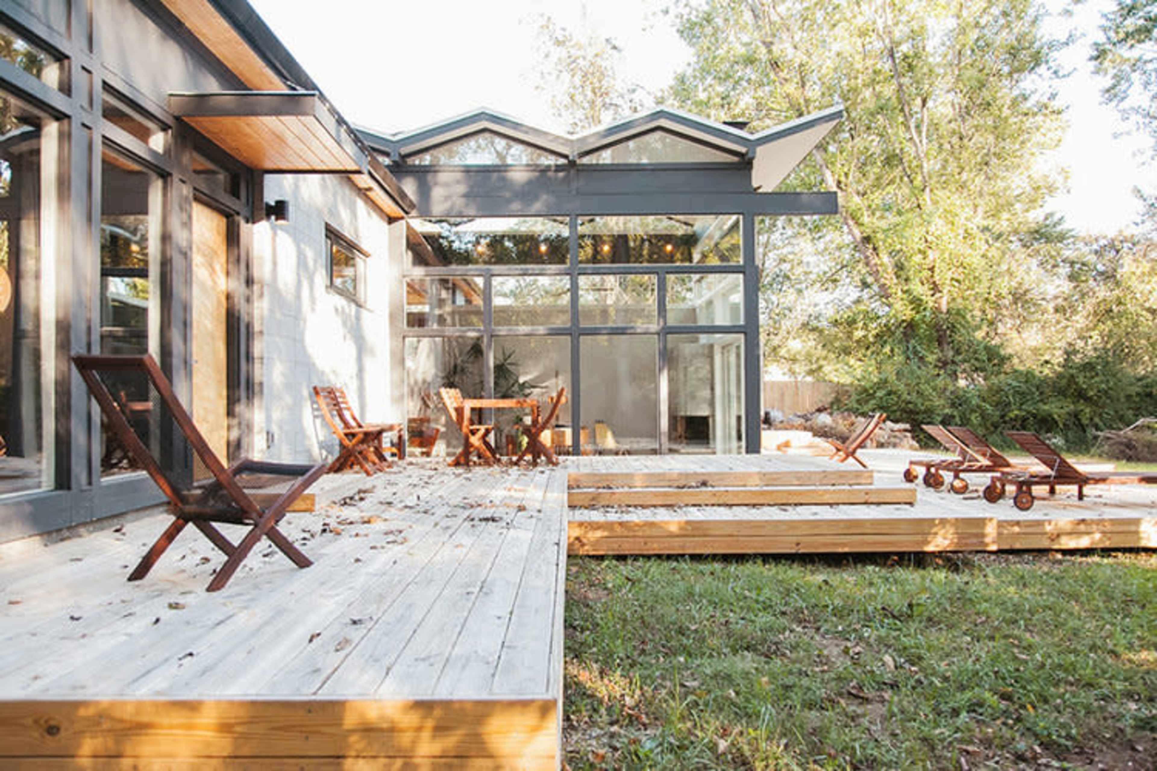 A modern home with large windows features a wooden deck and several outdoor seating areas in a forested setting.
