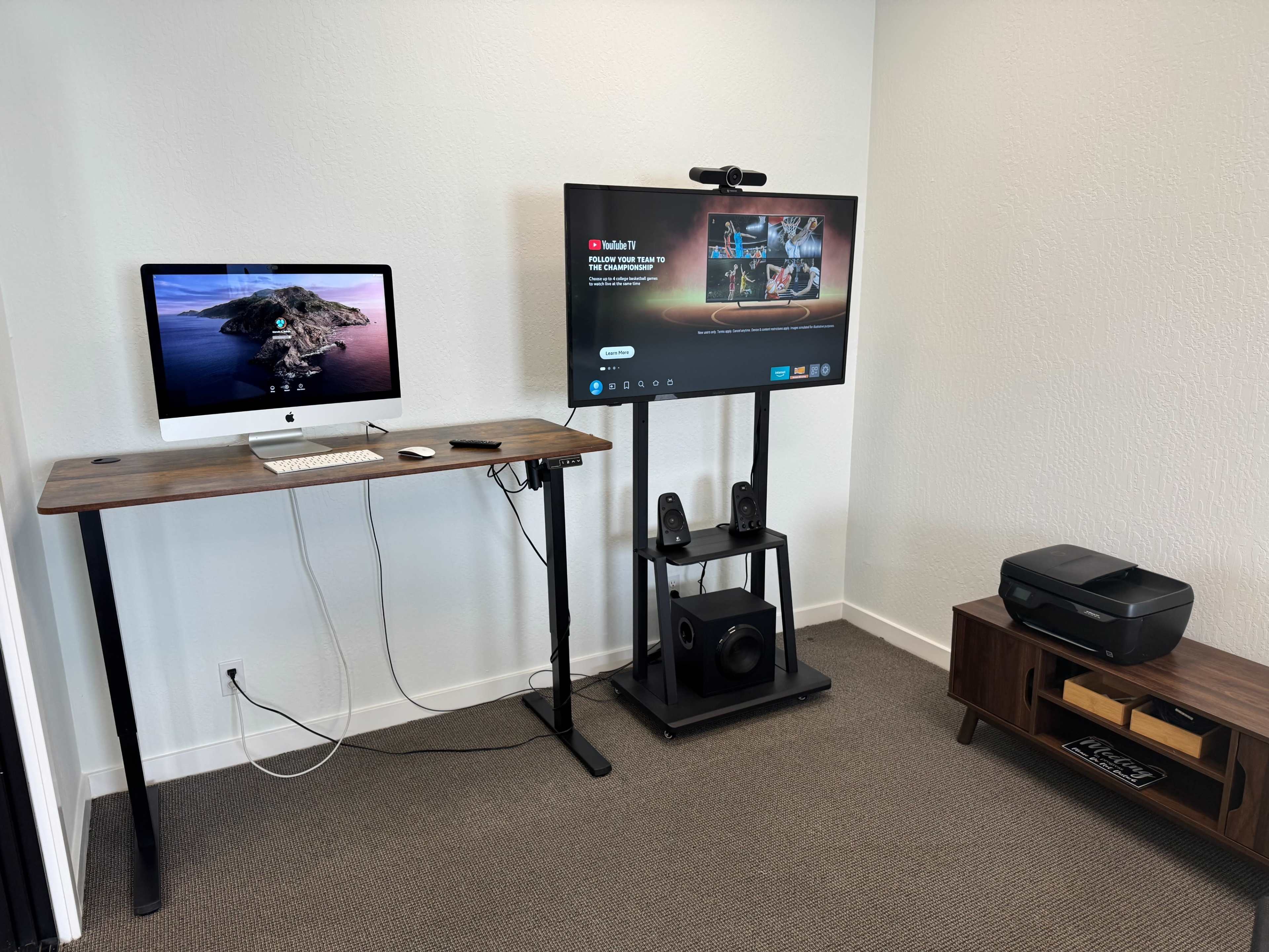 The image shows a modern workspace featuring an iMac on a wooden desk, a large screen mounted on a stand, speakers, and a printer on a nearby cabinet.