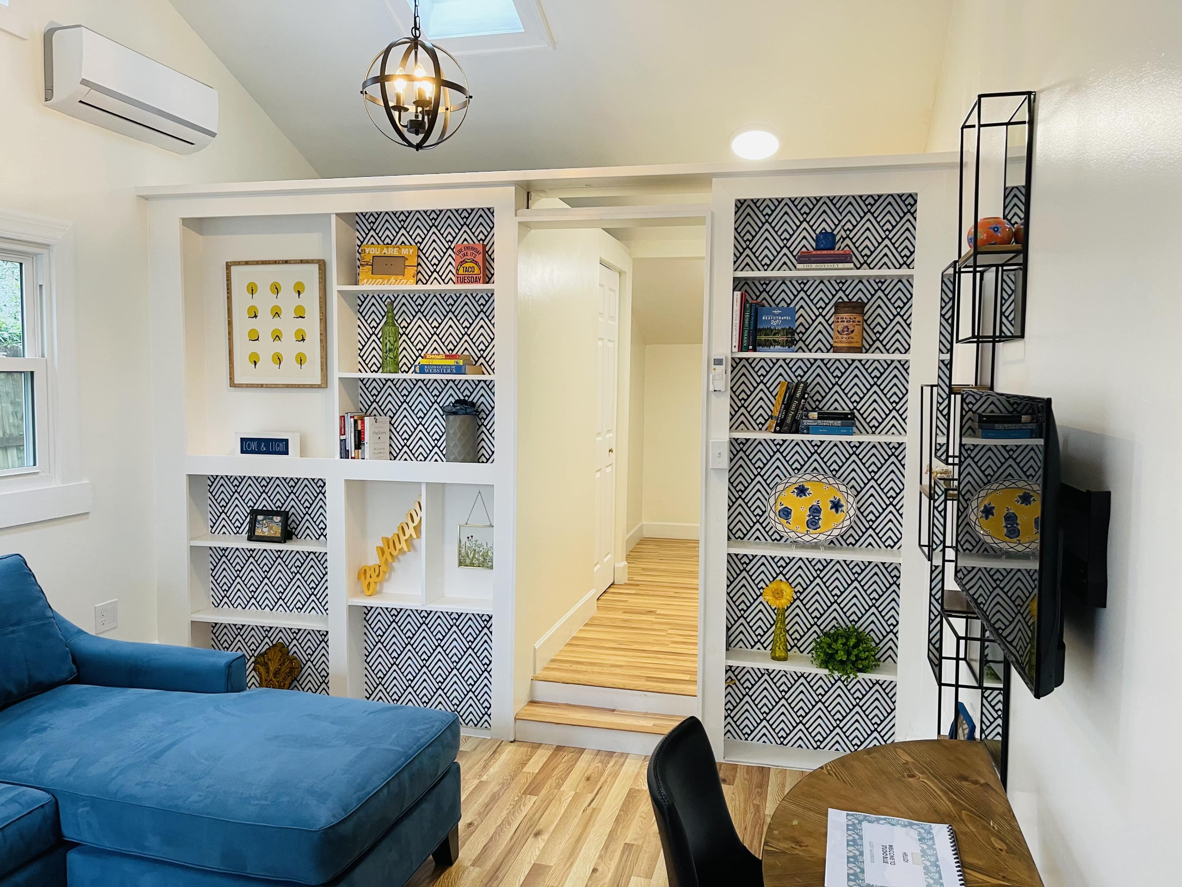 A modern, compact living space with a blue sofa, built-in shelves featuring decorative items, and a small workspace.