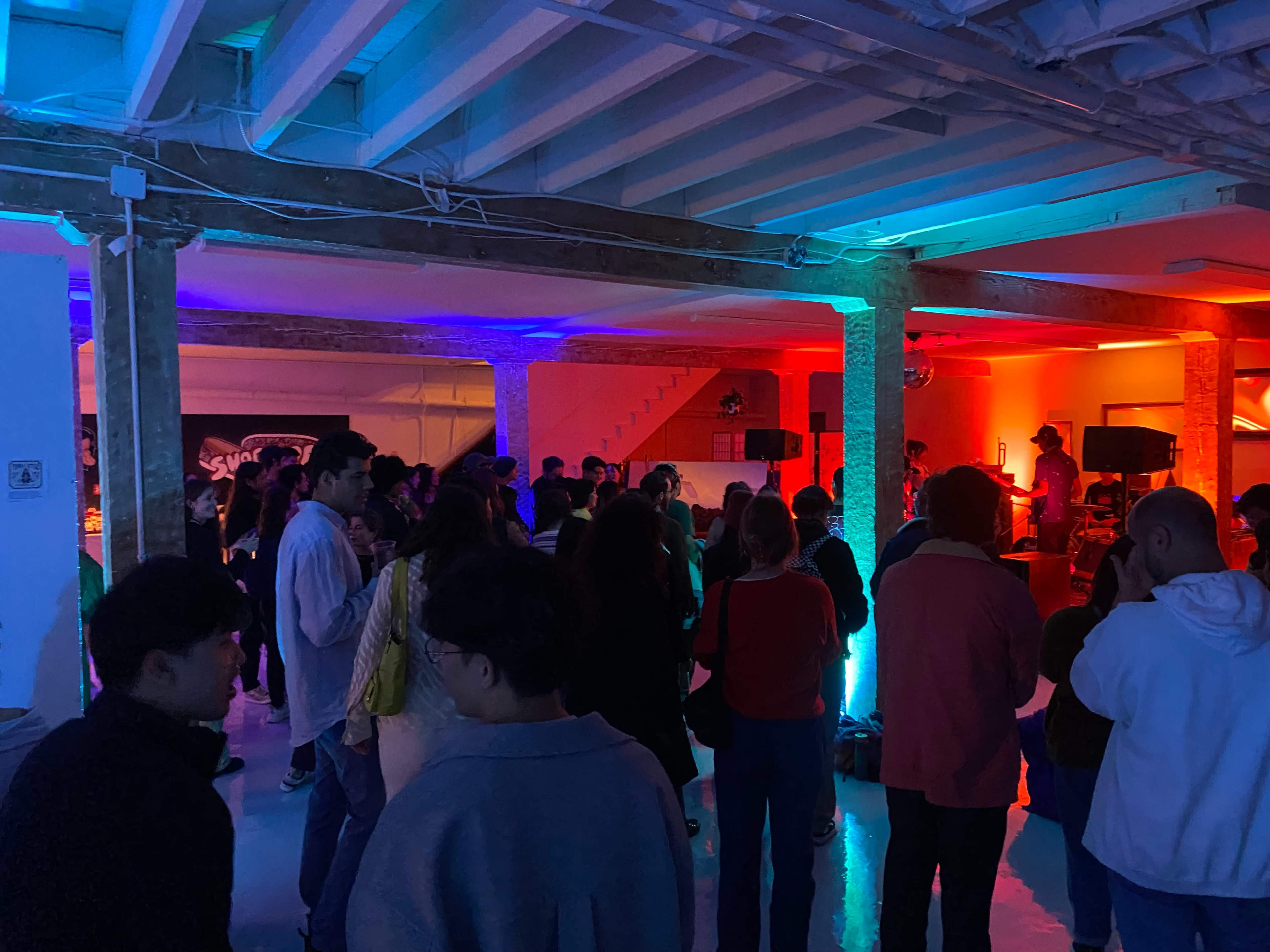 A crowded event space features people socializing and watching a live performance under colorful lighting.