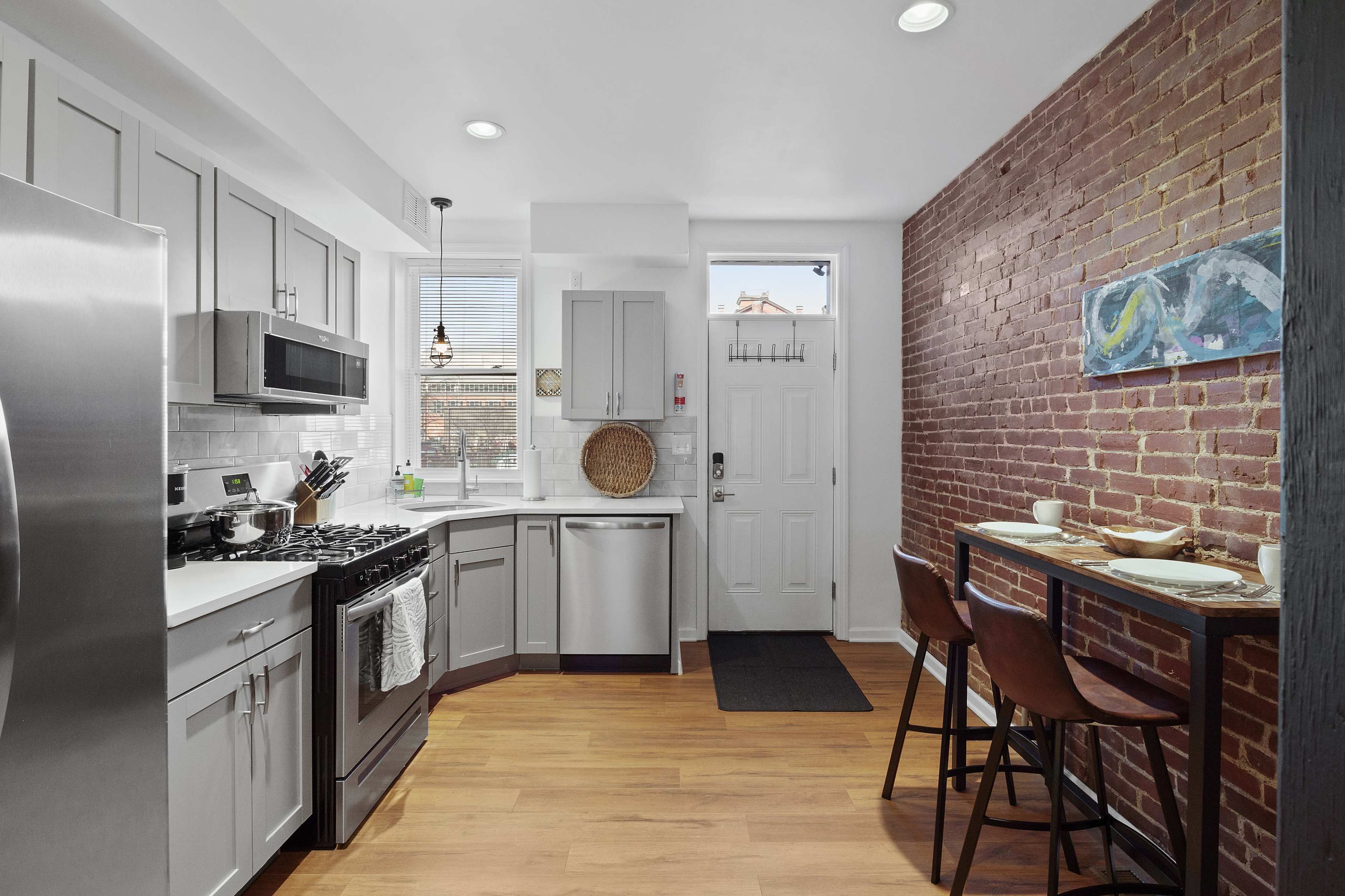 The image shows a modern kitchen with stainless steel appliances, gray cabinetry, and a brick accent wall, featuring a small dining area with two stools.