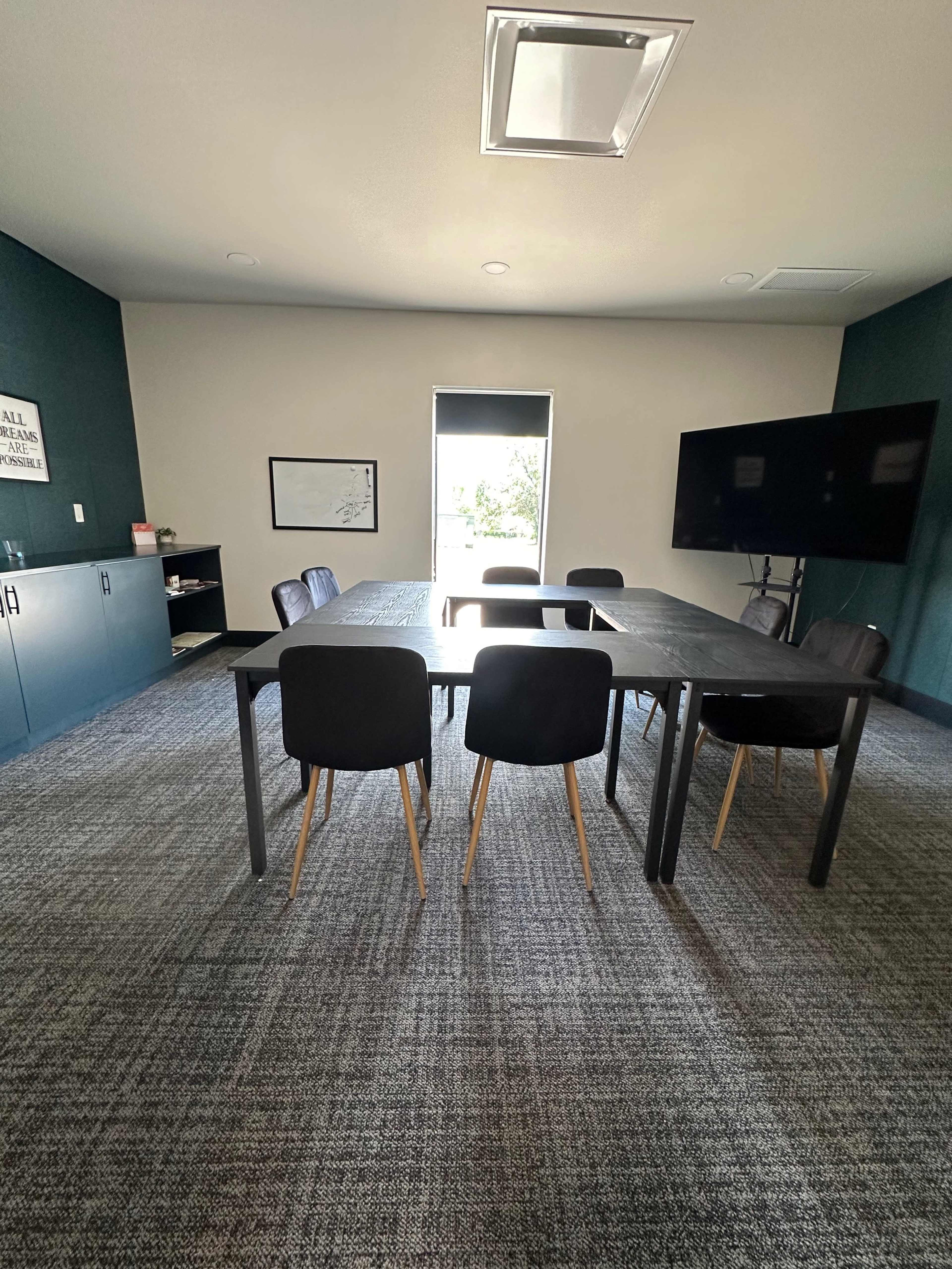 A conference room features a central table surrounded by six chairs, with a wall-mounted screen and a window allowing natural light.
