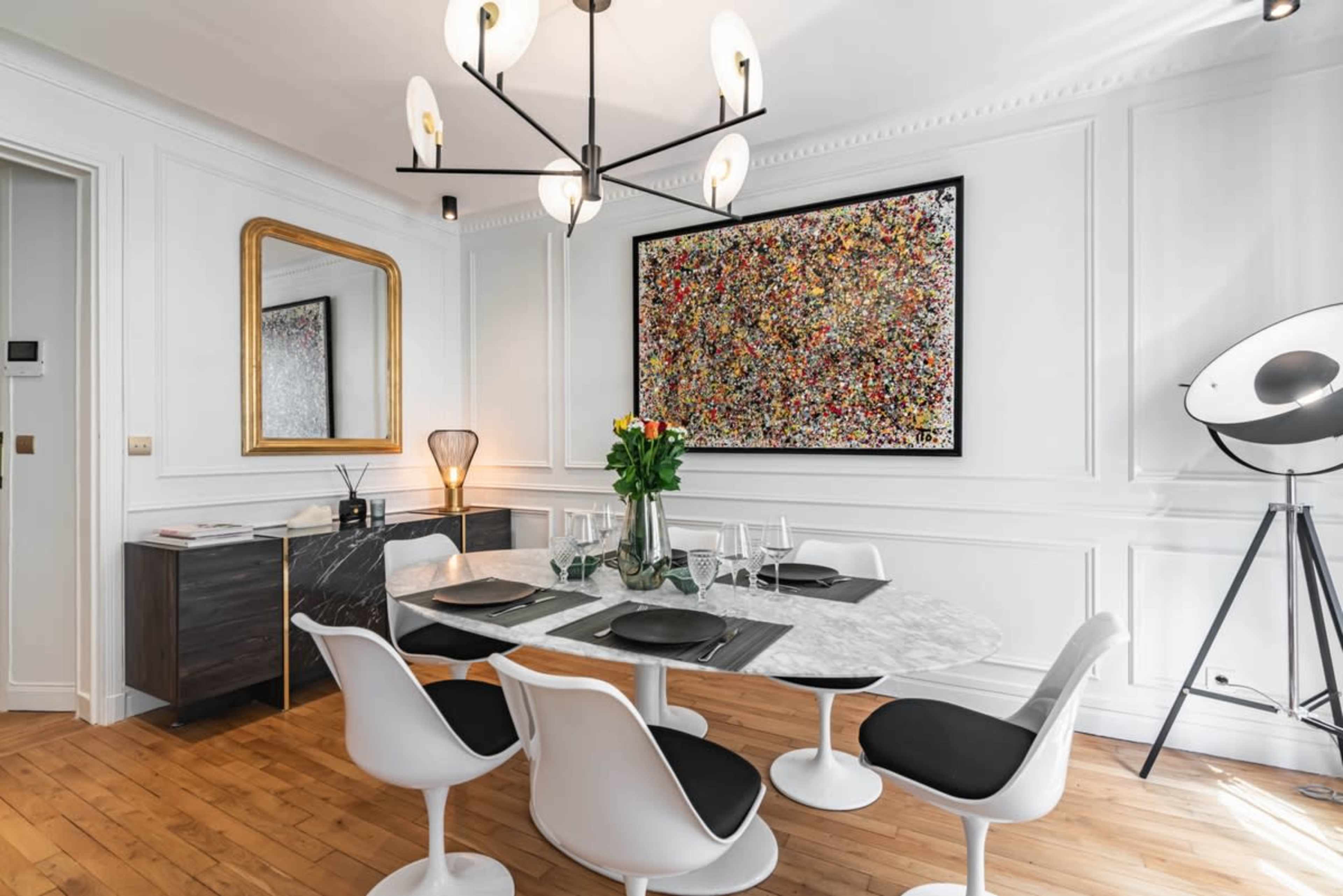 A modern dining area features a circular marble table set with black and white chairs, a colorful abstract painting on the wall, and contemporary lighting fixtures.