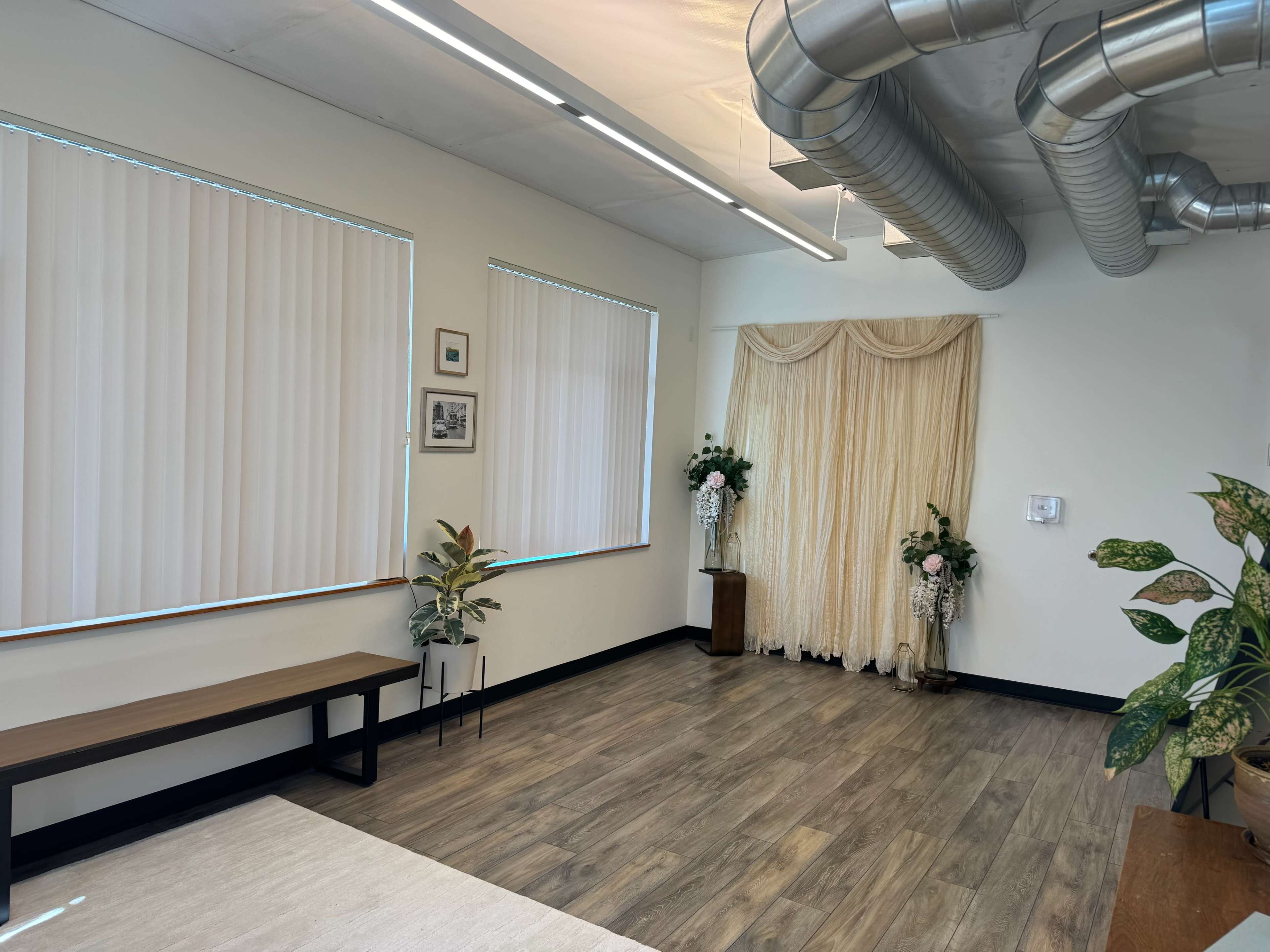 The image shows an interior space with wooden flooring, large windows covered by vertical blinds, a bench against one wall, and a floral arrangement near a draped curtain in the corner.