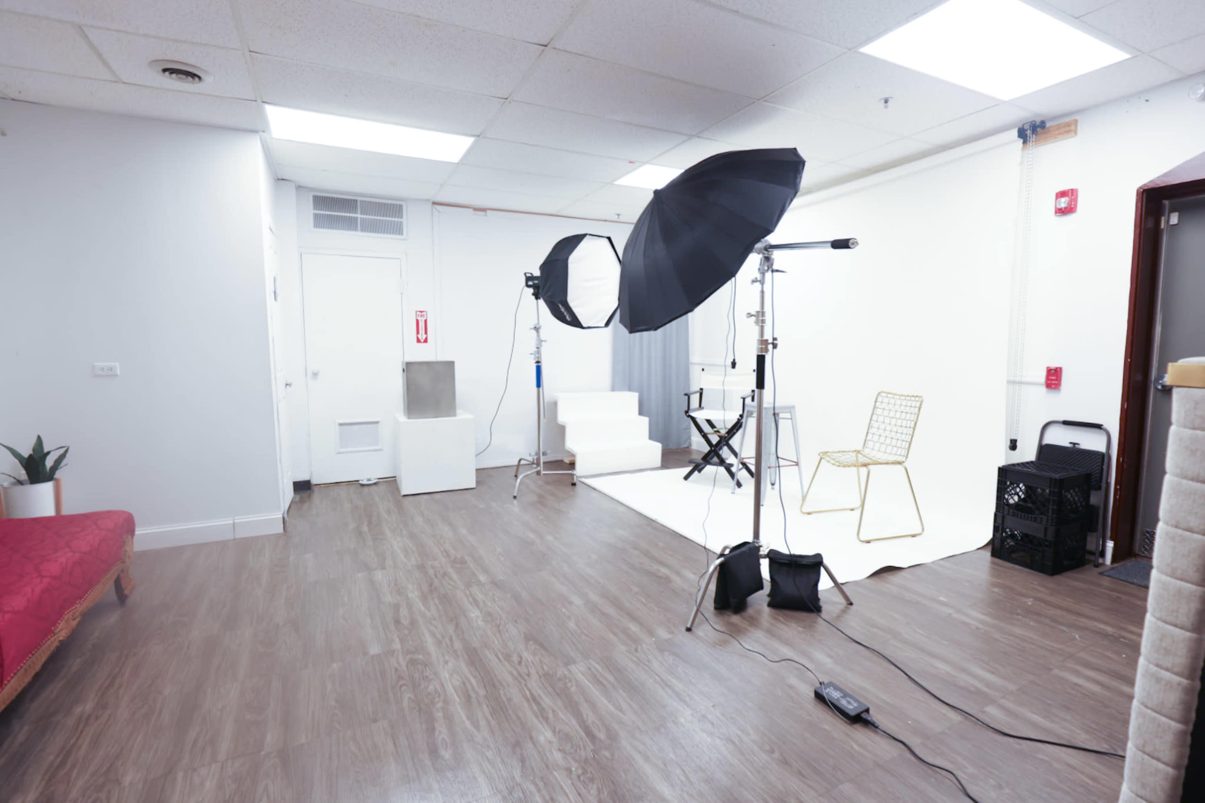A photography studio featuring white backdrop, lighting equipment, a chair, and various props arranged in a well-lit space.