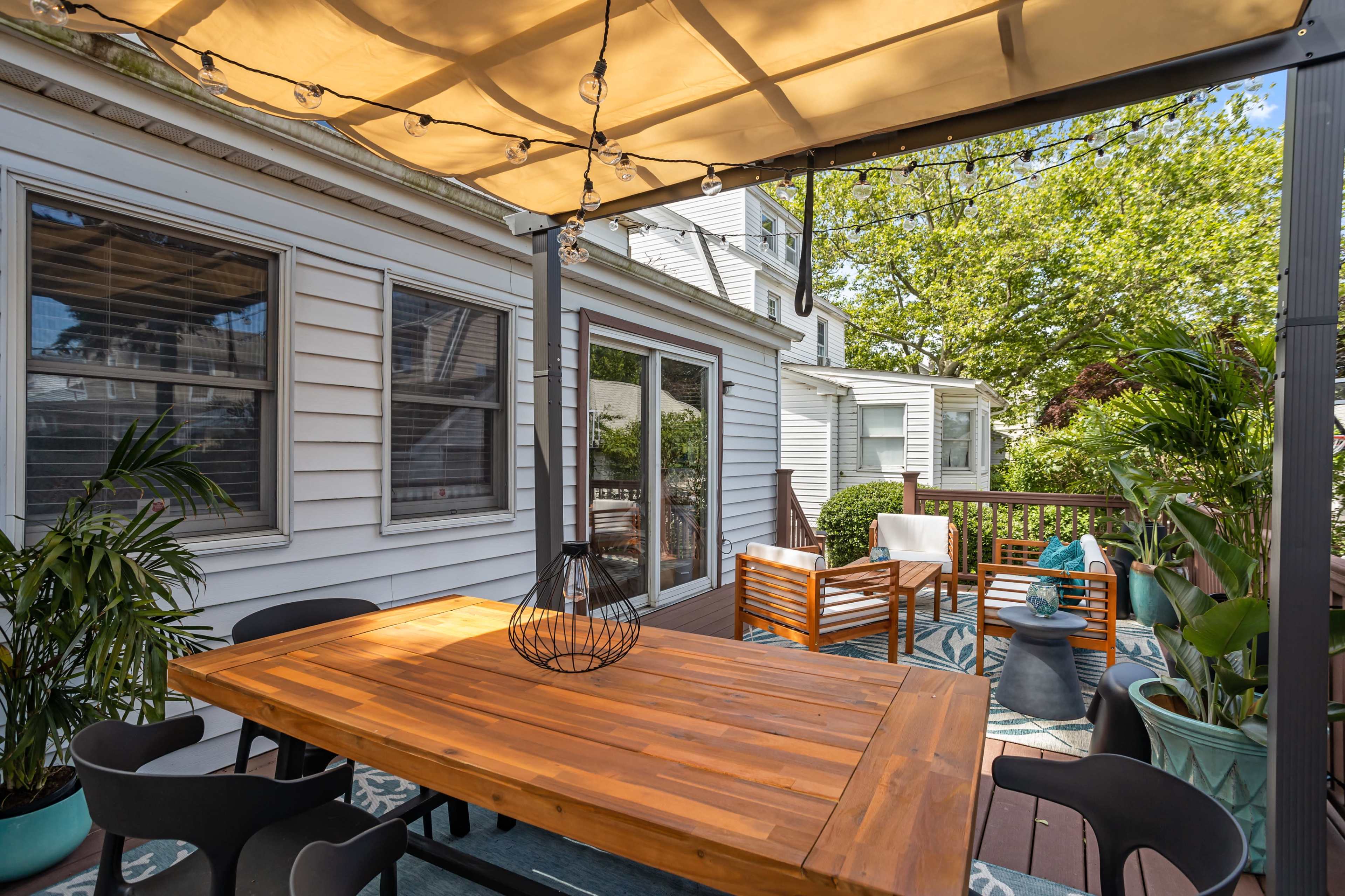 The image shows a spacious outdoor patio area featuring a wooden dining table, several seating arrangements, and potted plants under a canopy.