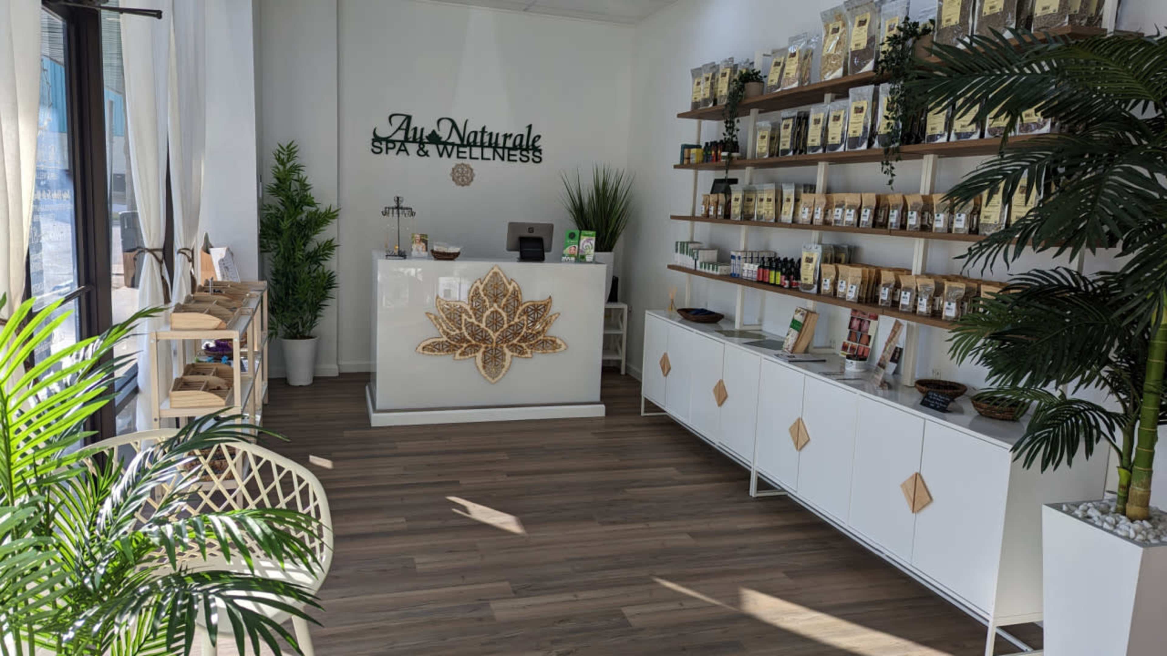 A spa and wellness reception area with wooden flooring, a front desk featuring a decorative lotus symbol, and shelves displaying various wellness products.