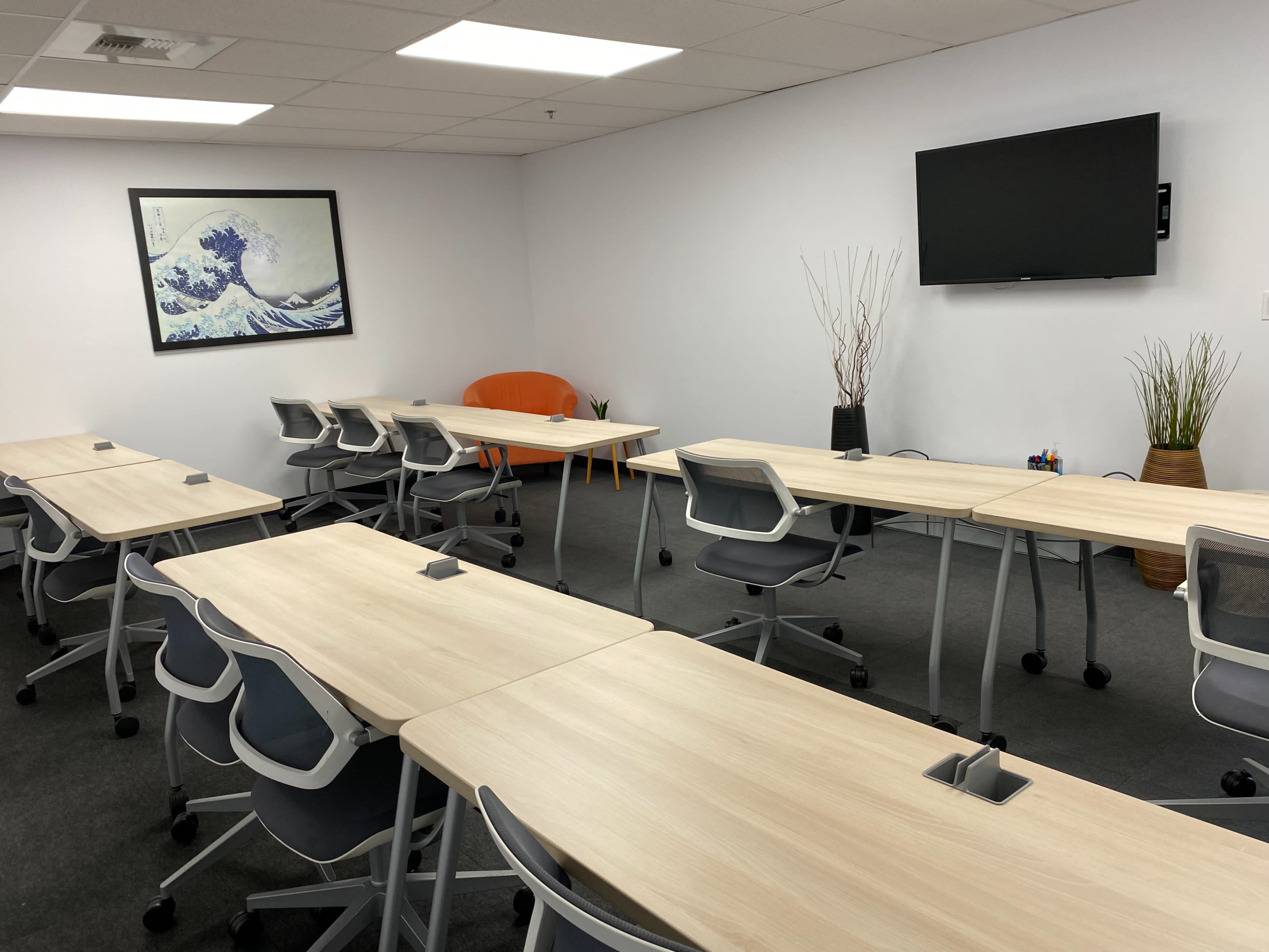 The image shows a modern classroom setup with multiple wooden tables and chairs arranged in rows, a television on the wall, and decorative items in the corners.