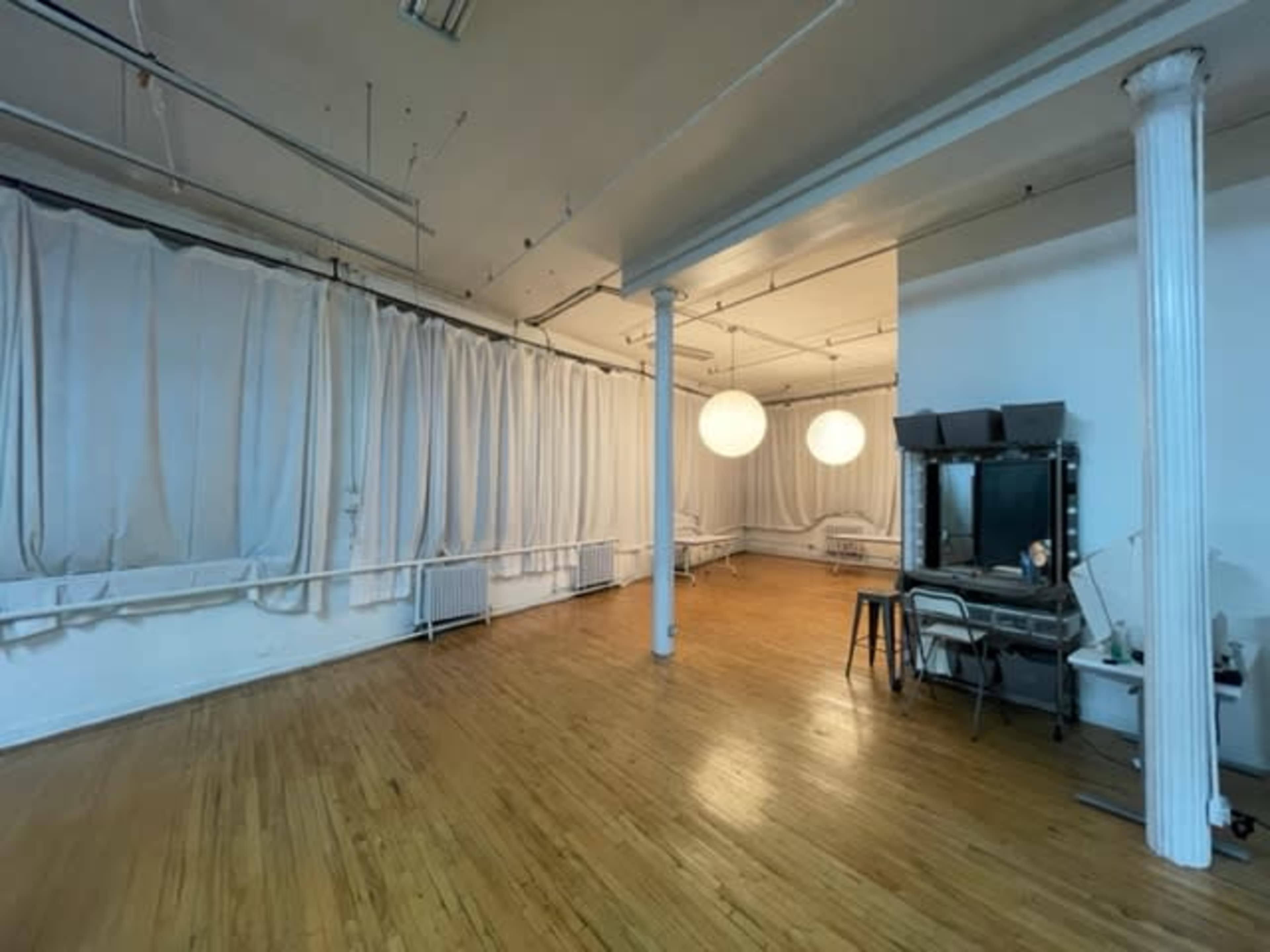 The image shows a spacious room with light wood flooring, white drapery on the walls, and two large round pendant lights hanging from the ceiling.