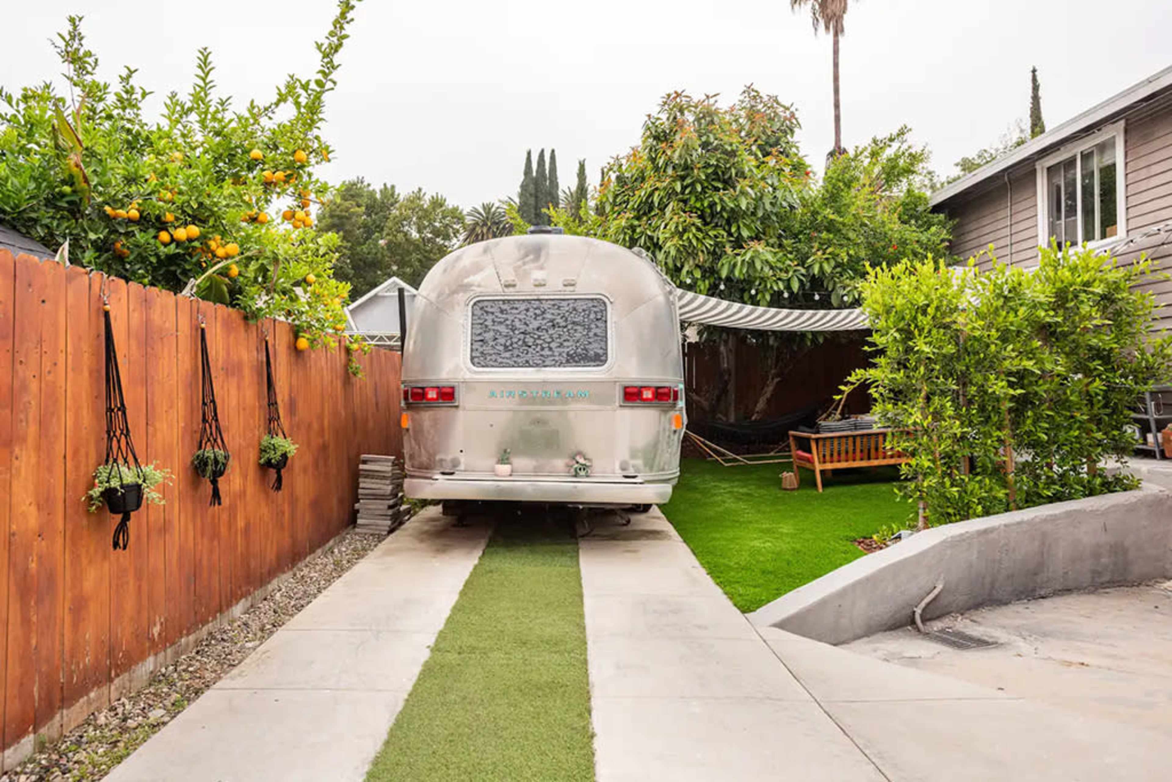 An Airstream trailer is parked between a wooden fence and a landscaped yard with greenery and a sitting area.