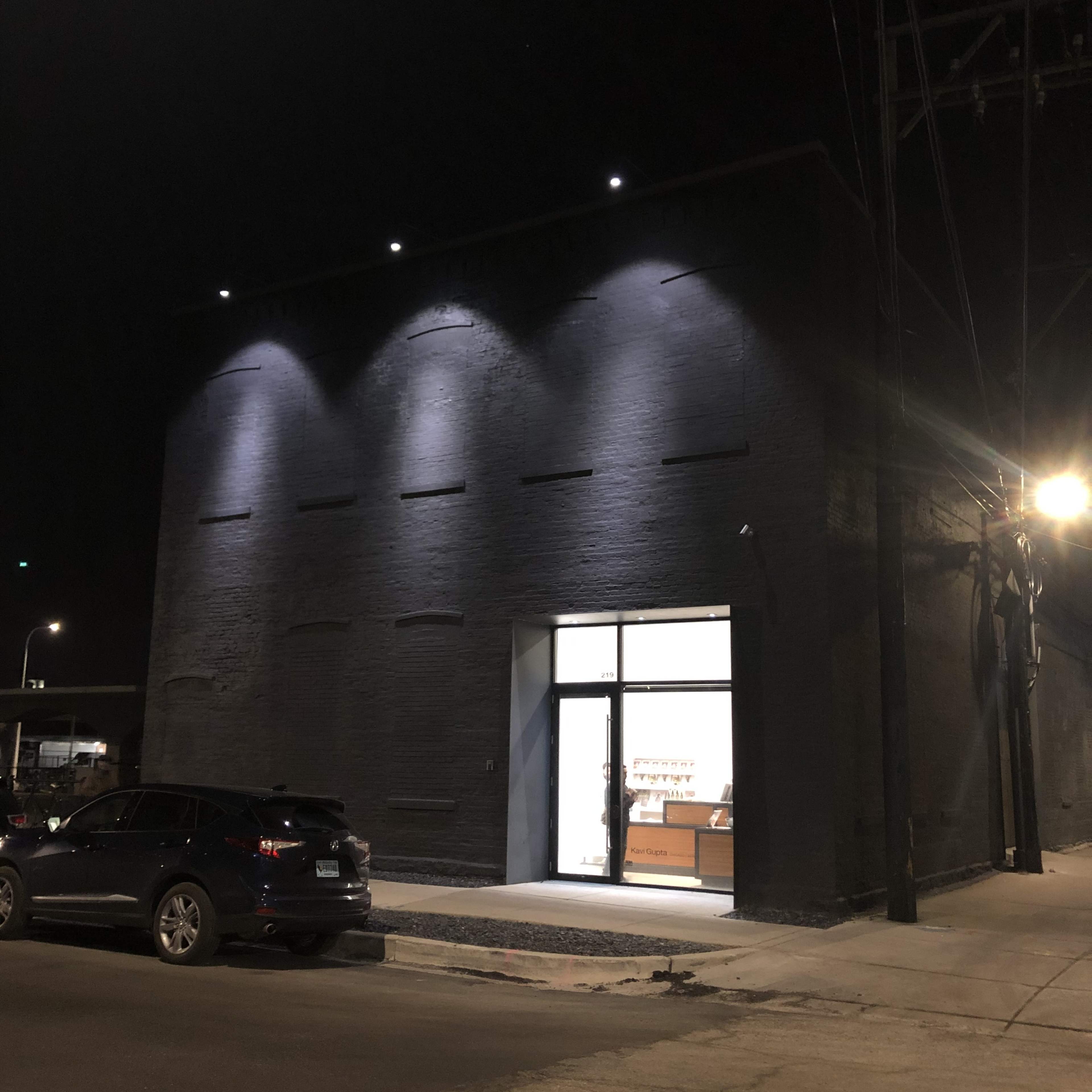 The image shows a dimly lit storefront with a large window displaying interior lighting, situated on a city street at night.