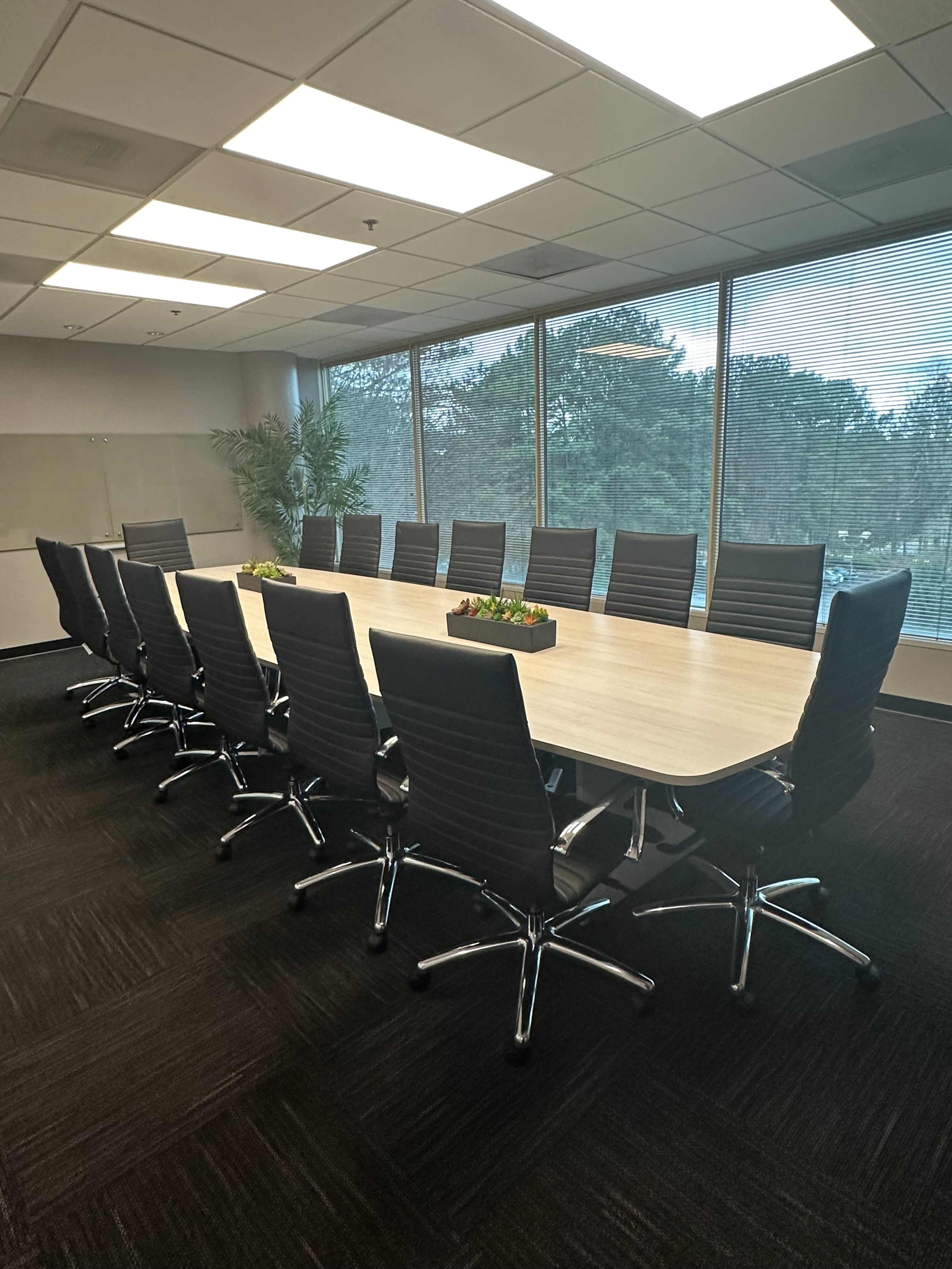 A long conference table surrounded by black swivel chairs is set up in a well-lit meeting room with large windows overlooking trees.