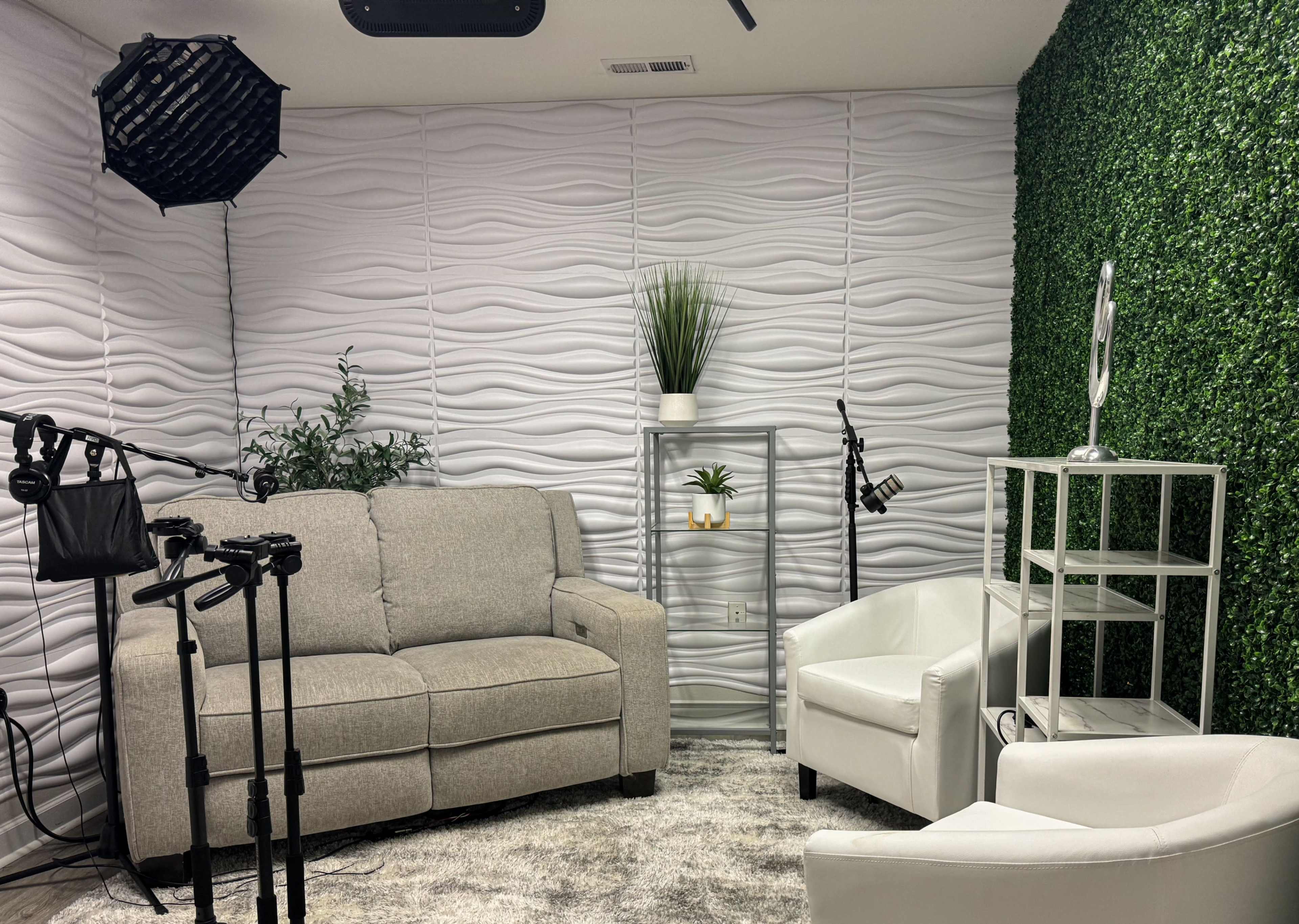 The image shows a modern indoor setting featuring a gray sofa, two white chairs, a decorative shelf with plants, and green wall paneling, all under a soft lighting arrangement.